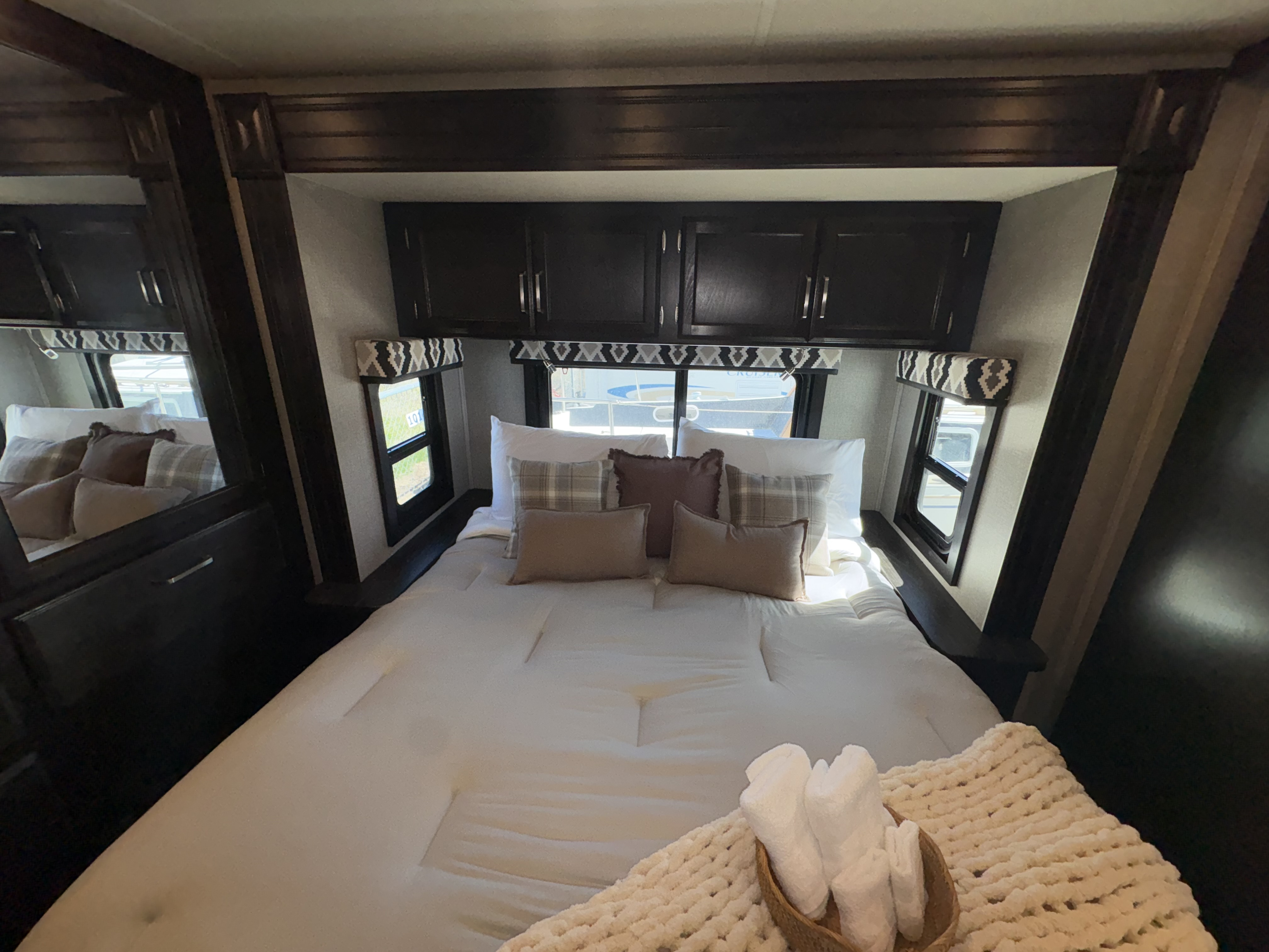 This rv rental bedroom features a comfy bed with white bedding, plaid pillows, dark cabinets, and side windows with patterned valances.. Jayco Greyhawk 2020