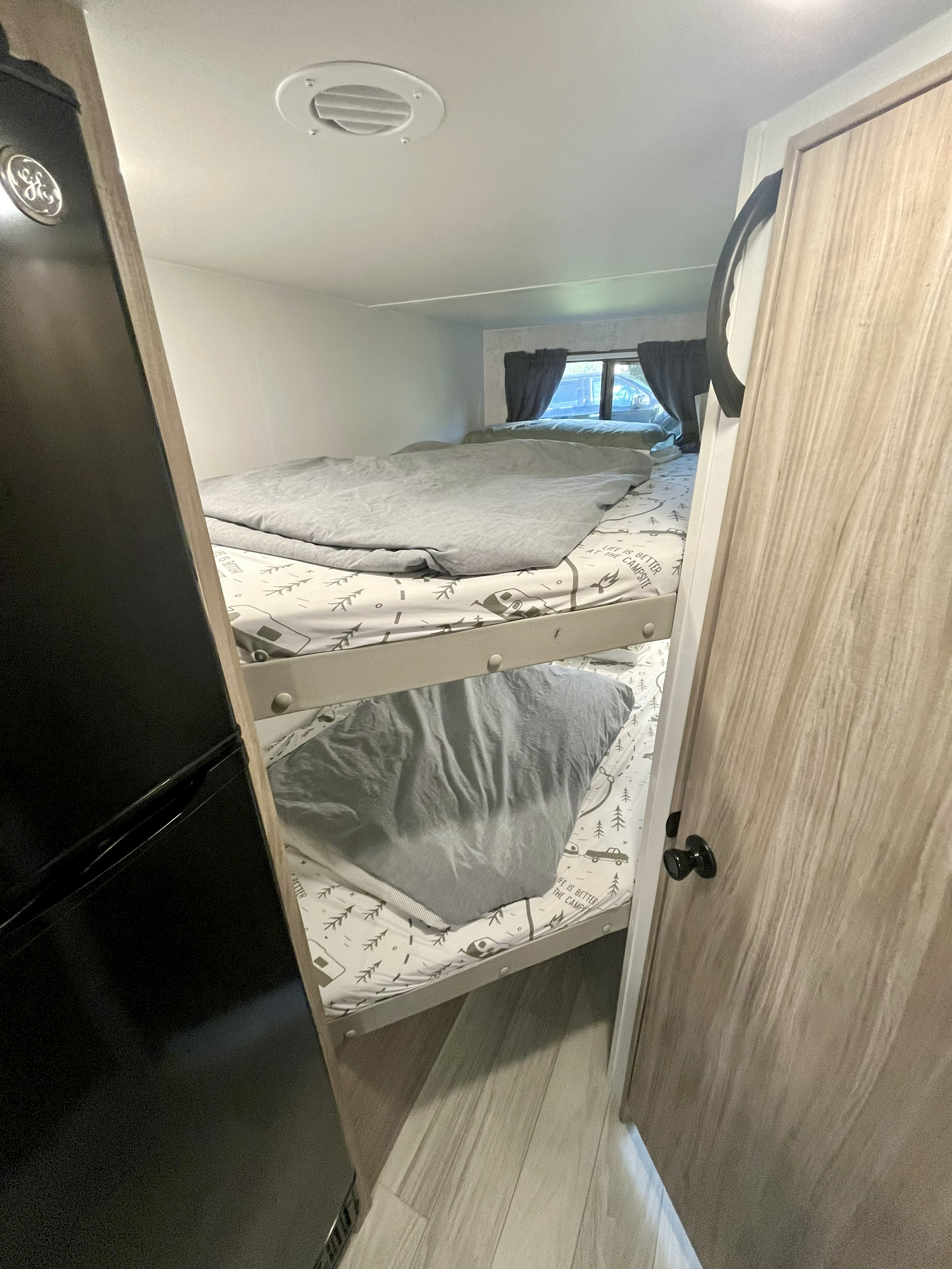 RV rental interior with stacked bunk beds, camping-patterned sheets, and a black refrigerator beside a wooden door.. Winnebago M Series 2025