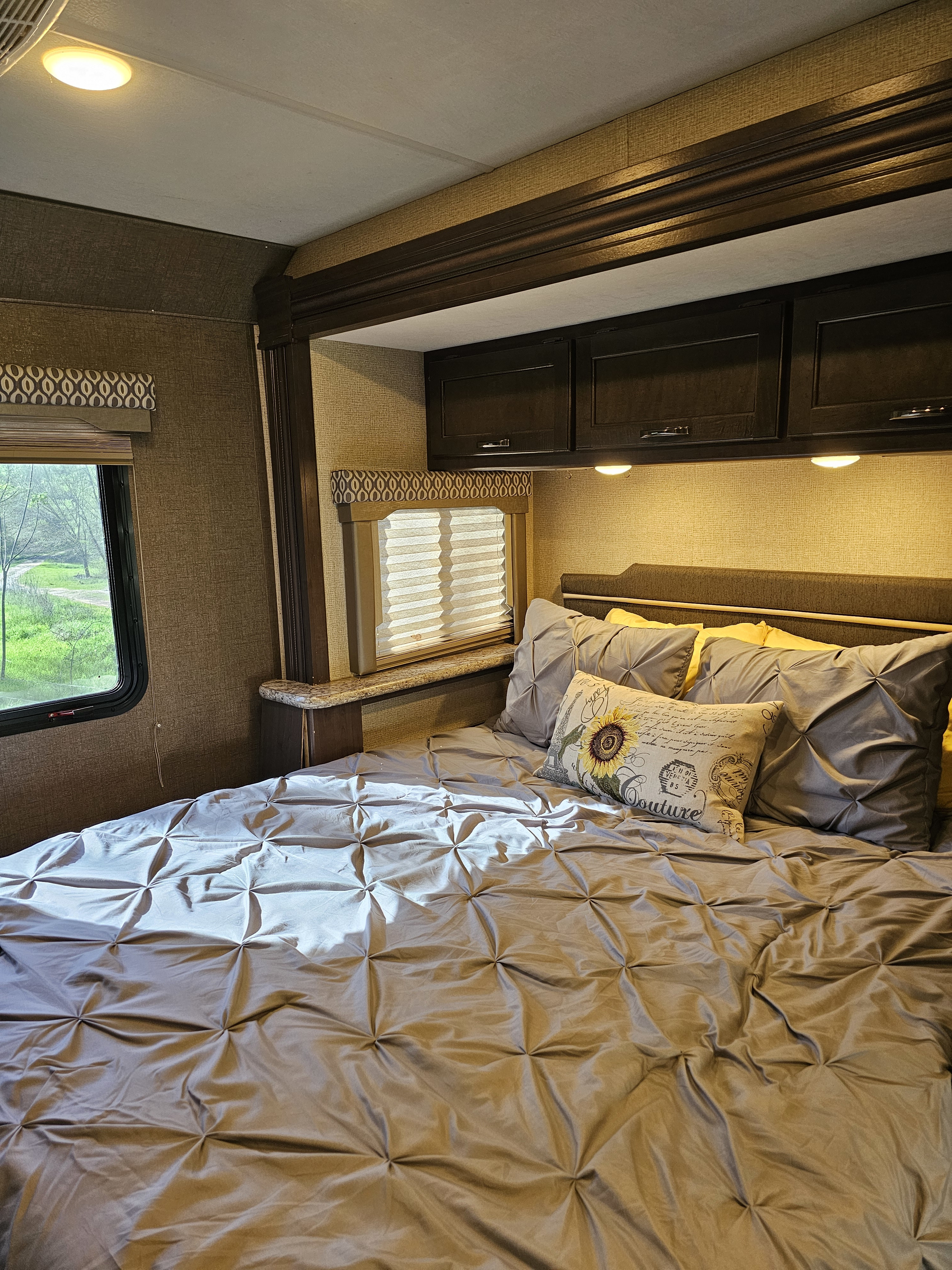 Cozy RV rental bedroom with a bed featuring gray and yellow pillows, overhead cabinets, and a window view.. Thor Freedom Elite 2018