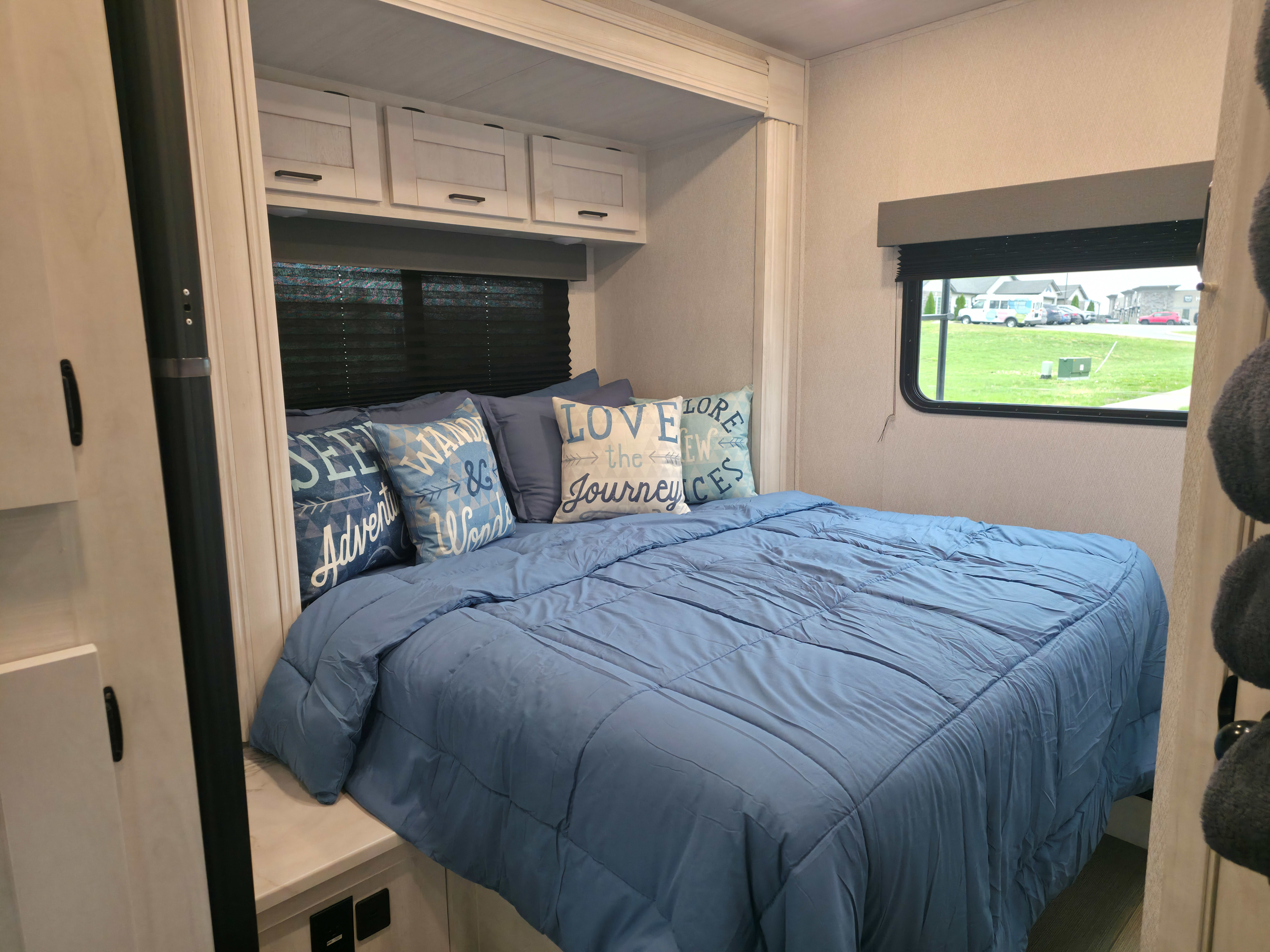 Cozy RV rental bedroom with blue bedding, inspirational pillows, overhead storage, and a window view, perfect for your next adventure.. East to West Entrada 2024