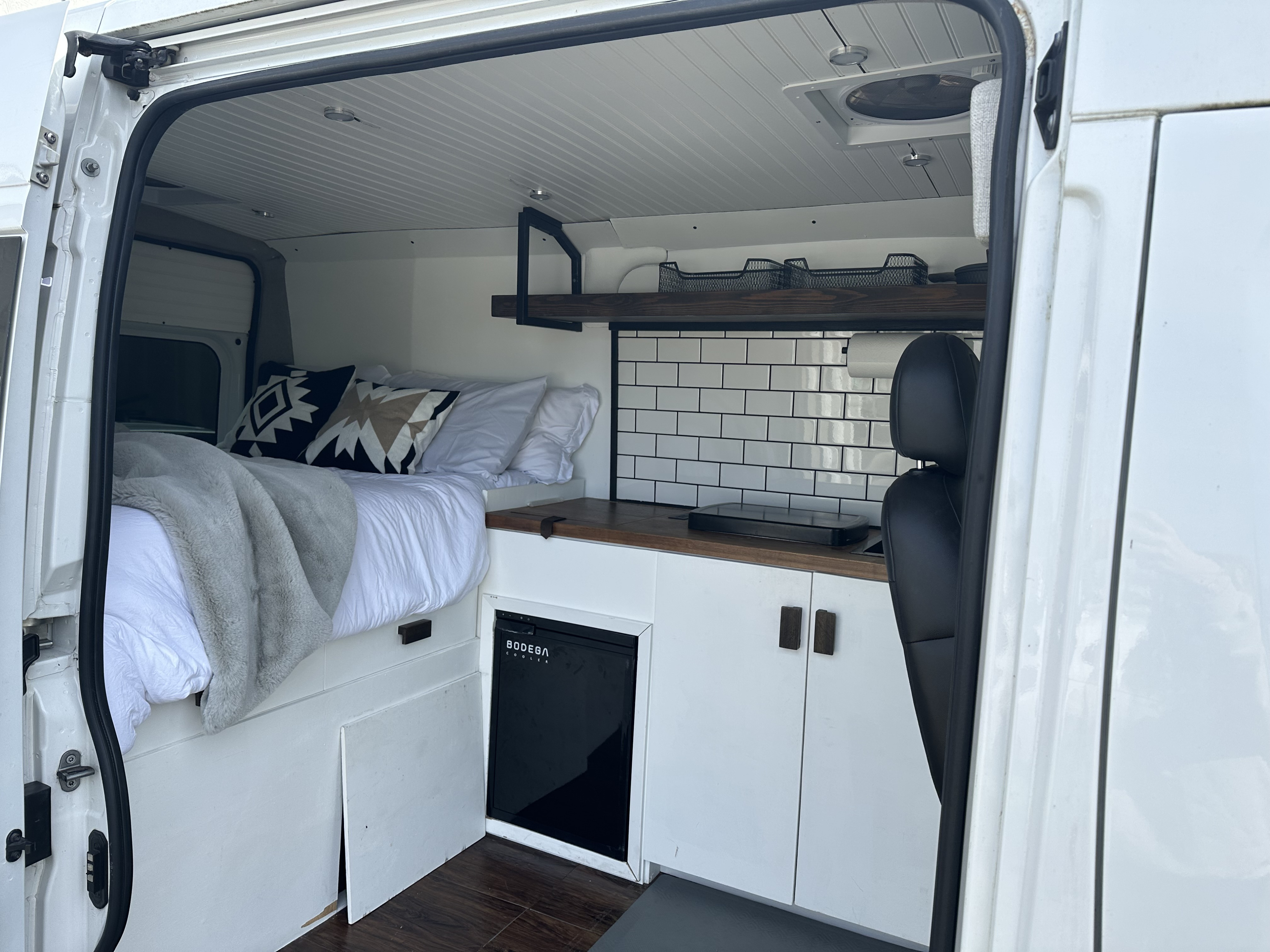 Cozy camper van interior for RV rental: bed, kitchen counter, tiled backsplash, cooler, and white cabinets.. Dodge Ram 1500 2016