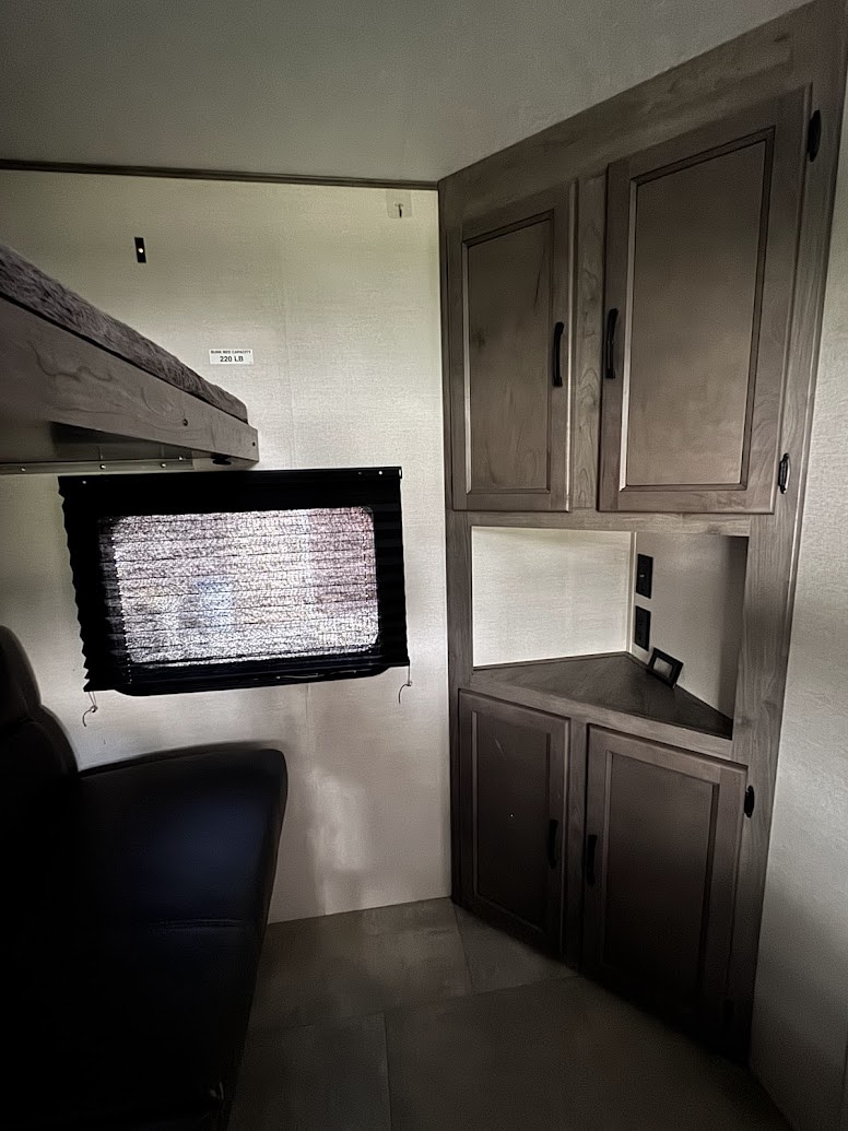 Cozy RV rental interior featuring a bunk bed, window with blind, and a functional corner cabinet unit with storage and outlets.. Coachmen Apex 2021