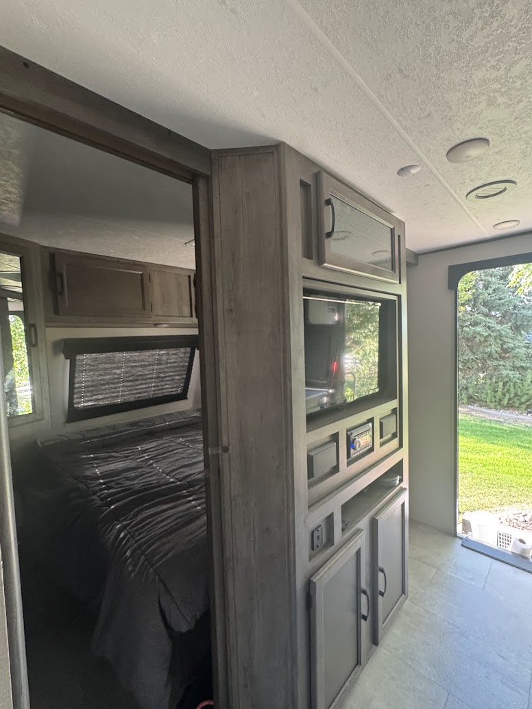 Modern RV rental interior showing a cozy bedroom with gray bedding and a built-in entertainment unit with a TV and storage.. Coachmen Apex 2021