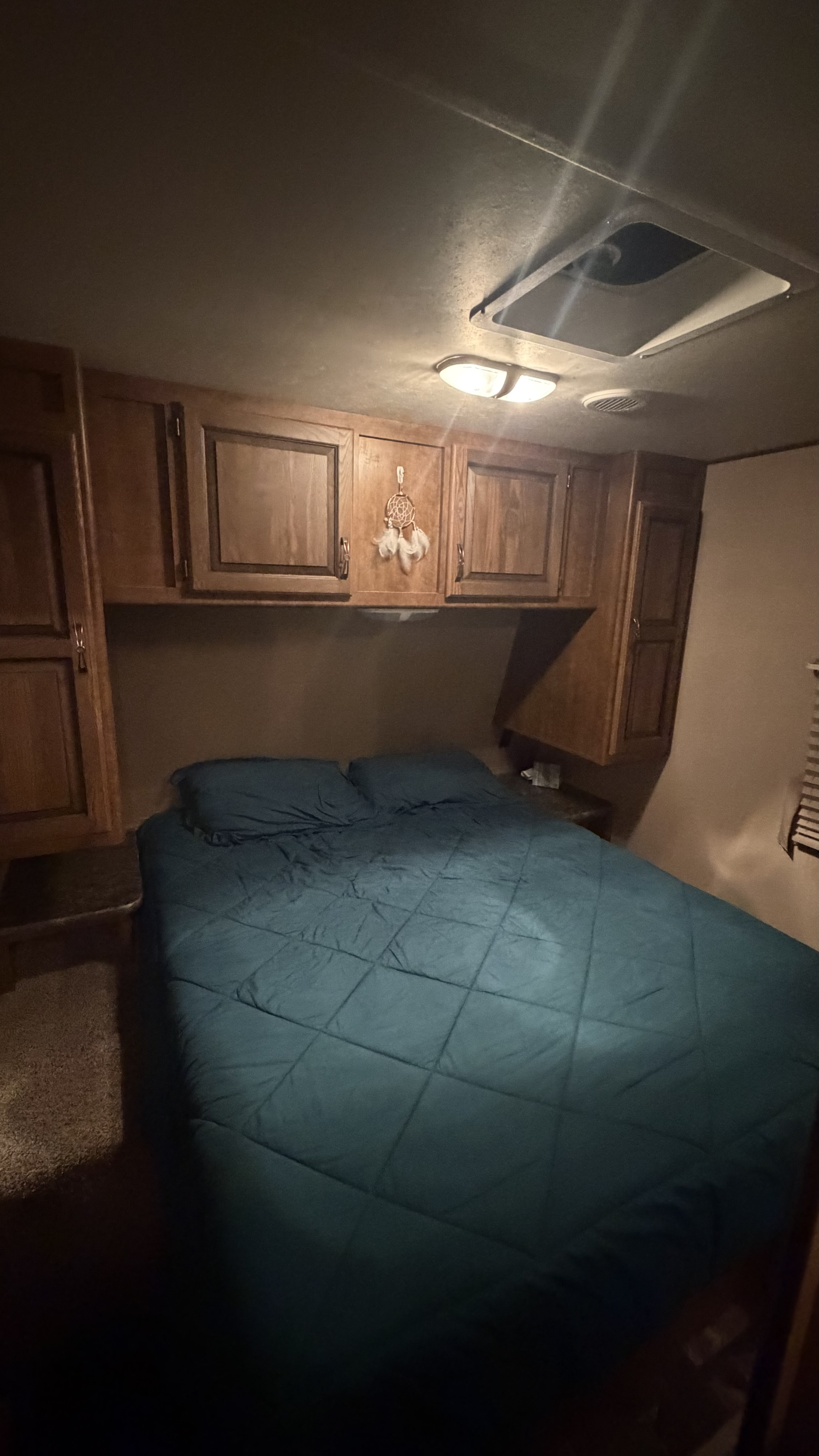 Compact RV rental bedroom with a dark blue bed, wooden overhead cabinets, and ceiling vent for a comfortable stay.. Crossroads RV Rezerve 2015