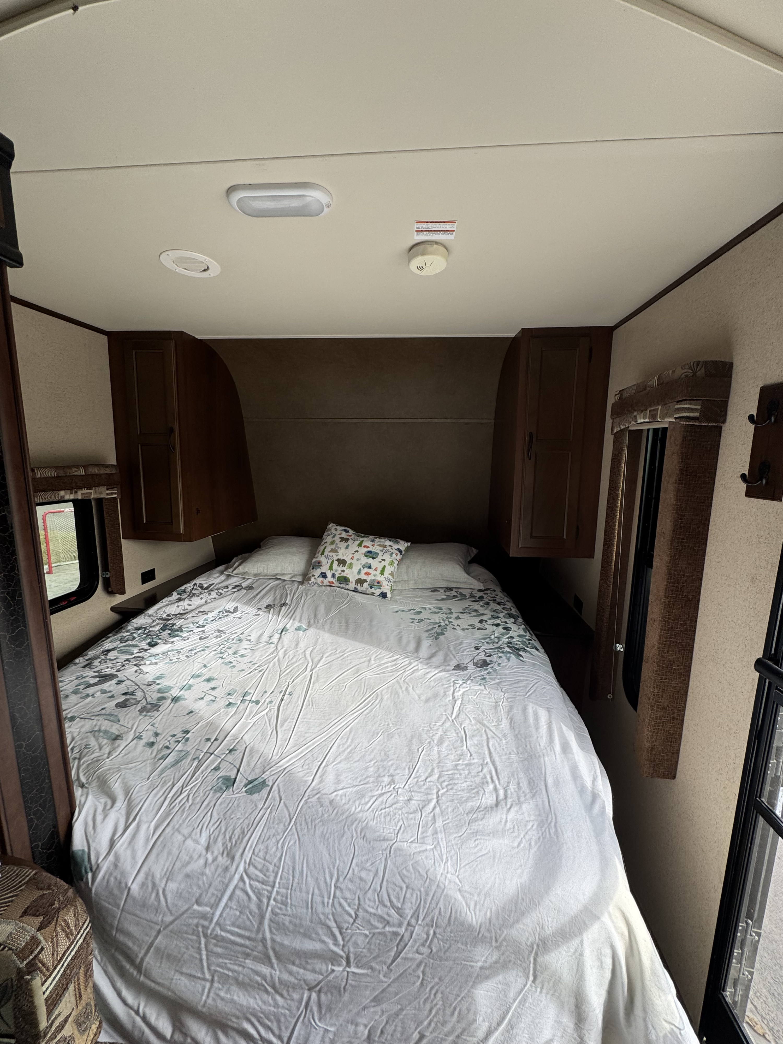 Compact RV rental bedroom with a bed featuring white patterned bedding, overhead cabinets, and side windows.. Jayco Jay Flight 2015