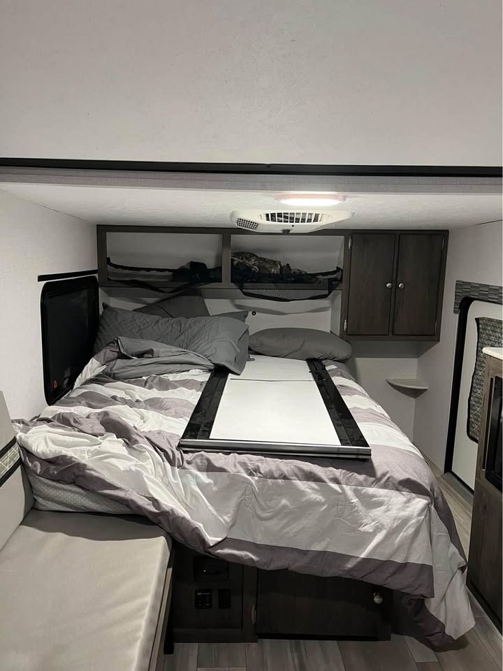 This cozy RV rental bedroom features striped bedding, overhead storage, an AC vent, and a small bench for comfort.. Forest River V-Tec Lifted Camper & Toy Hauler 2023