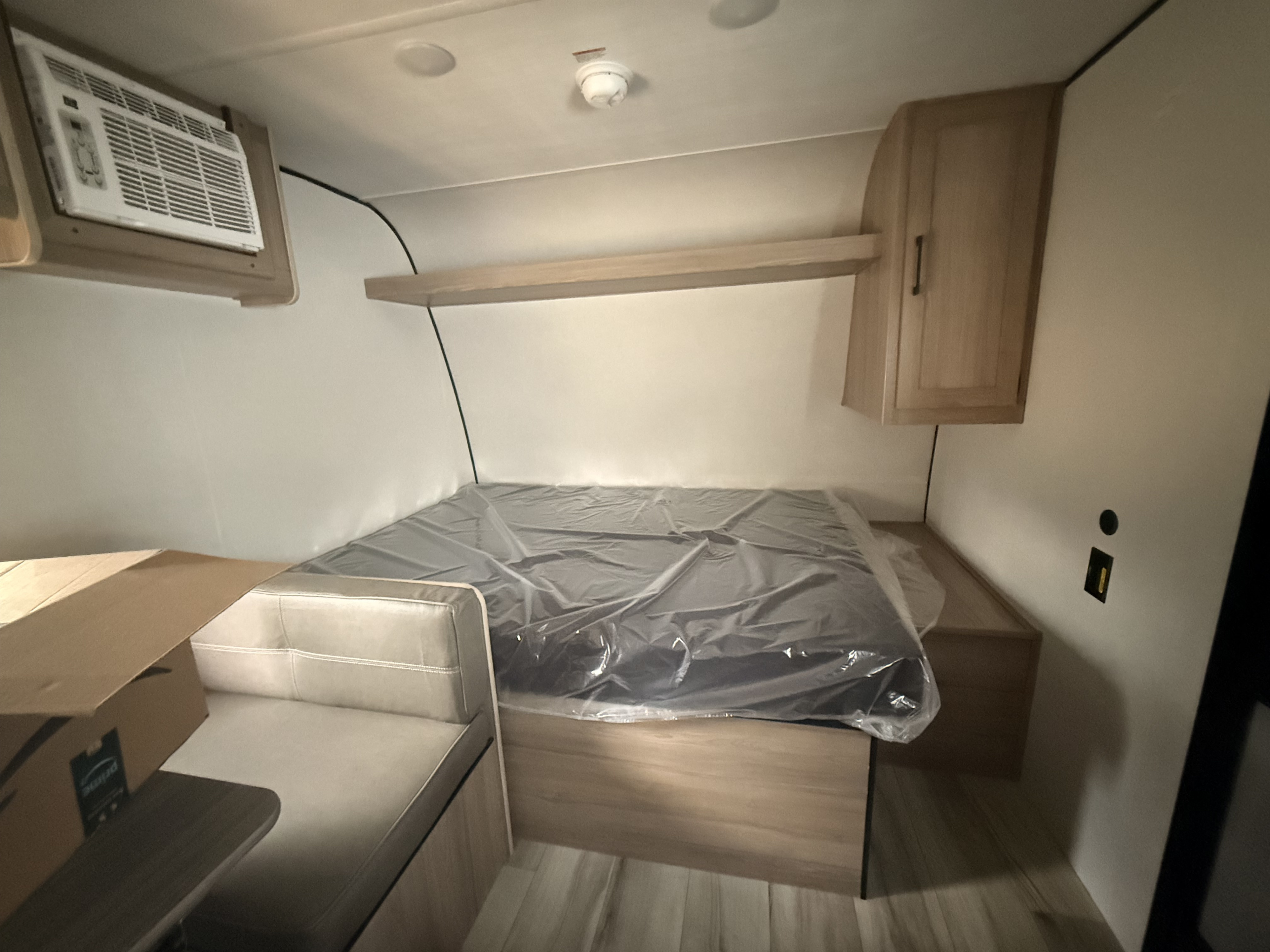 Bright RV rental interior with a bed in protective plastic, dinette bench, wall AC, overhead shelf, and cabinet.. Keystone Coleman 17B 2026