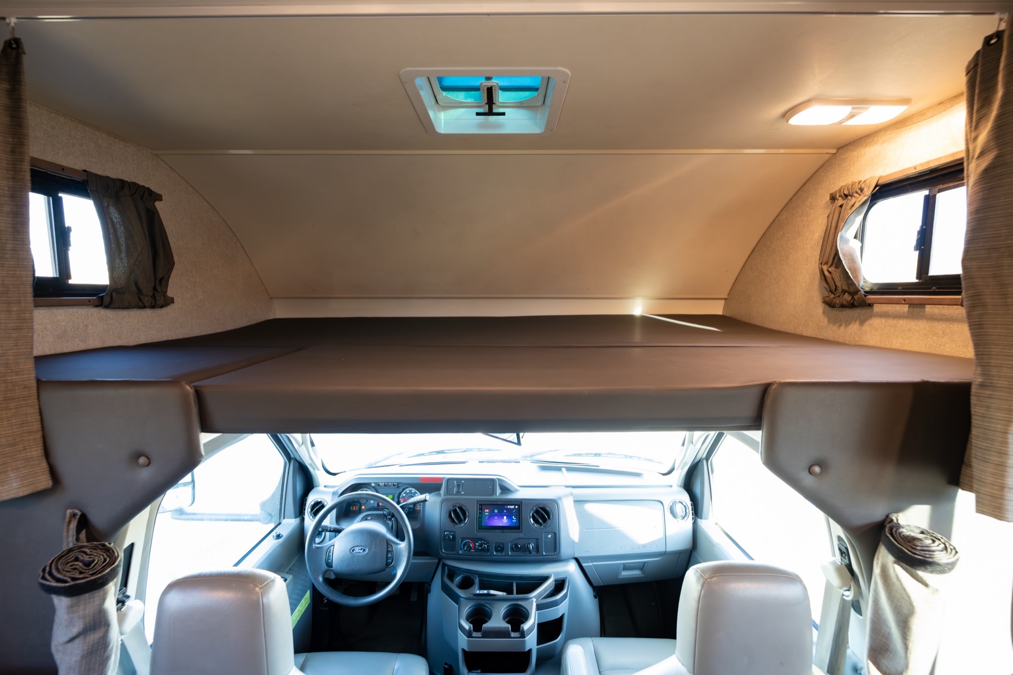Inside an rv rental: an overhead bunk bed extends above the driver's cabin, with a steering wheel and dashboard visible below.. Winnebago Minnie Winnie 2020