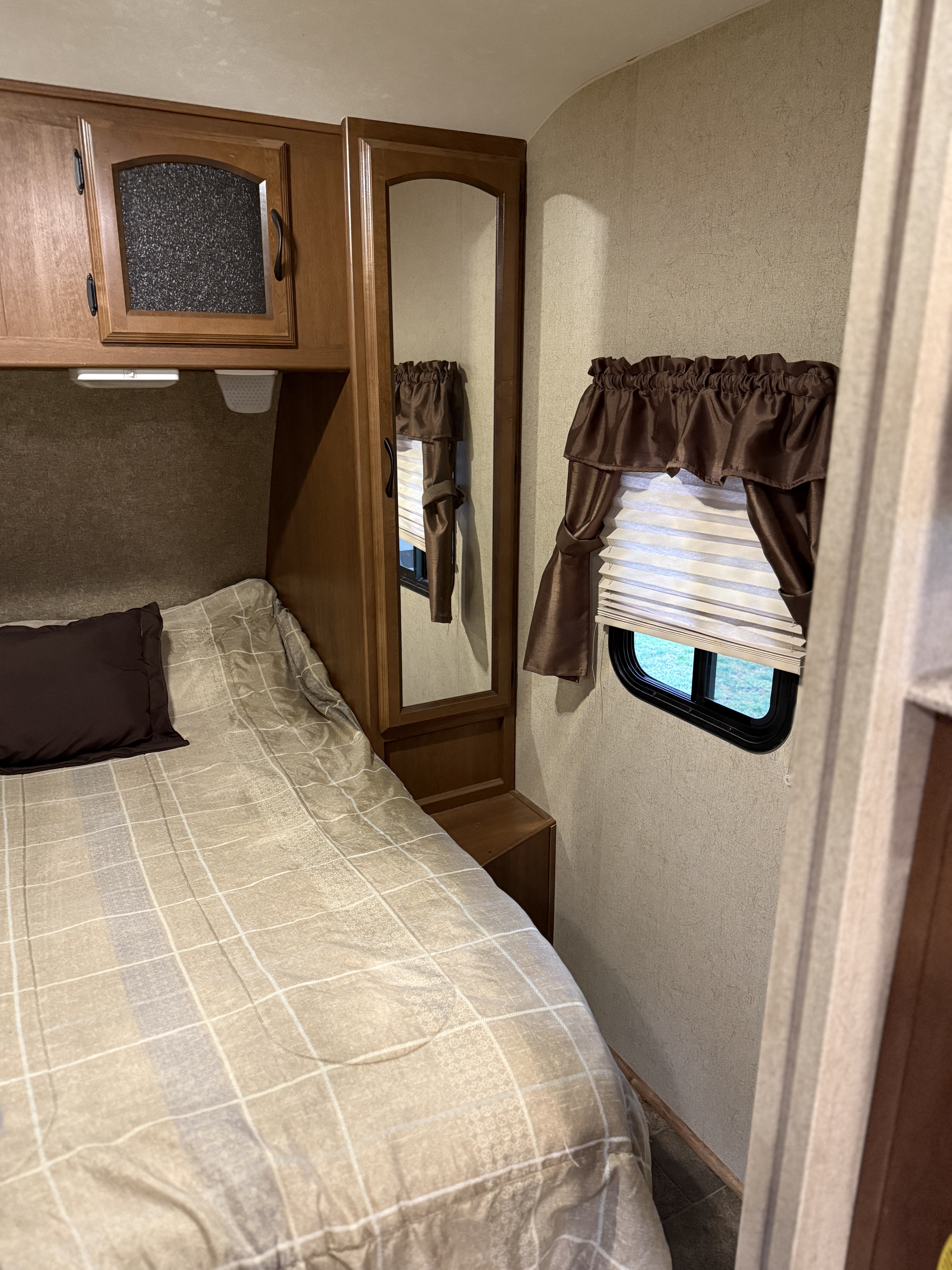 A cozy RV rental bedroom features a patterned bed, wooden cabinets with a full-length mirror, and a window with brown curtains.. Forest River Coachmen Freedom Express 2015