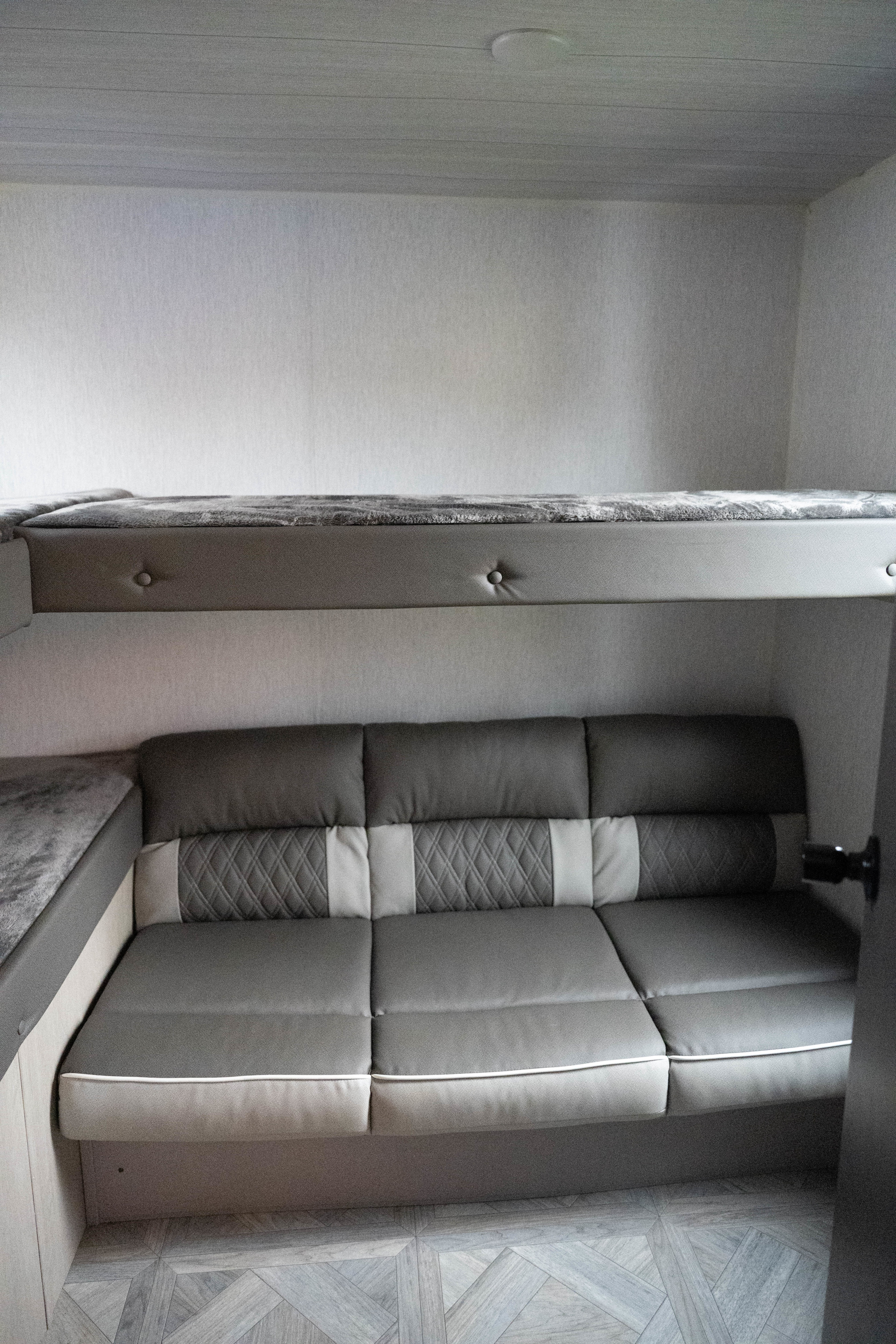 This RV rental interior features a modern gray sofa with quilted accents and an overhead bunk bed, ideal for compact living.. Forest River Salem Cruise Lite 2023