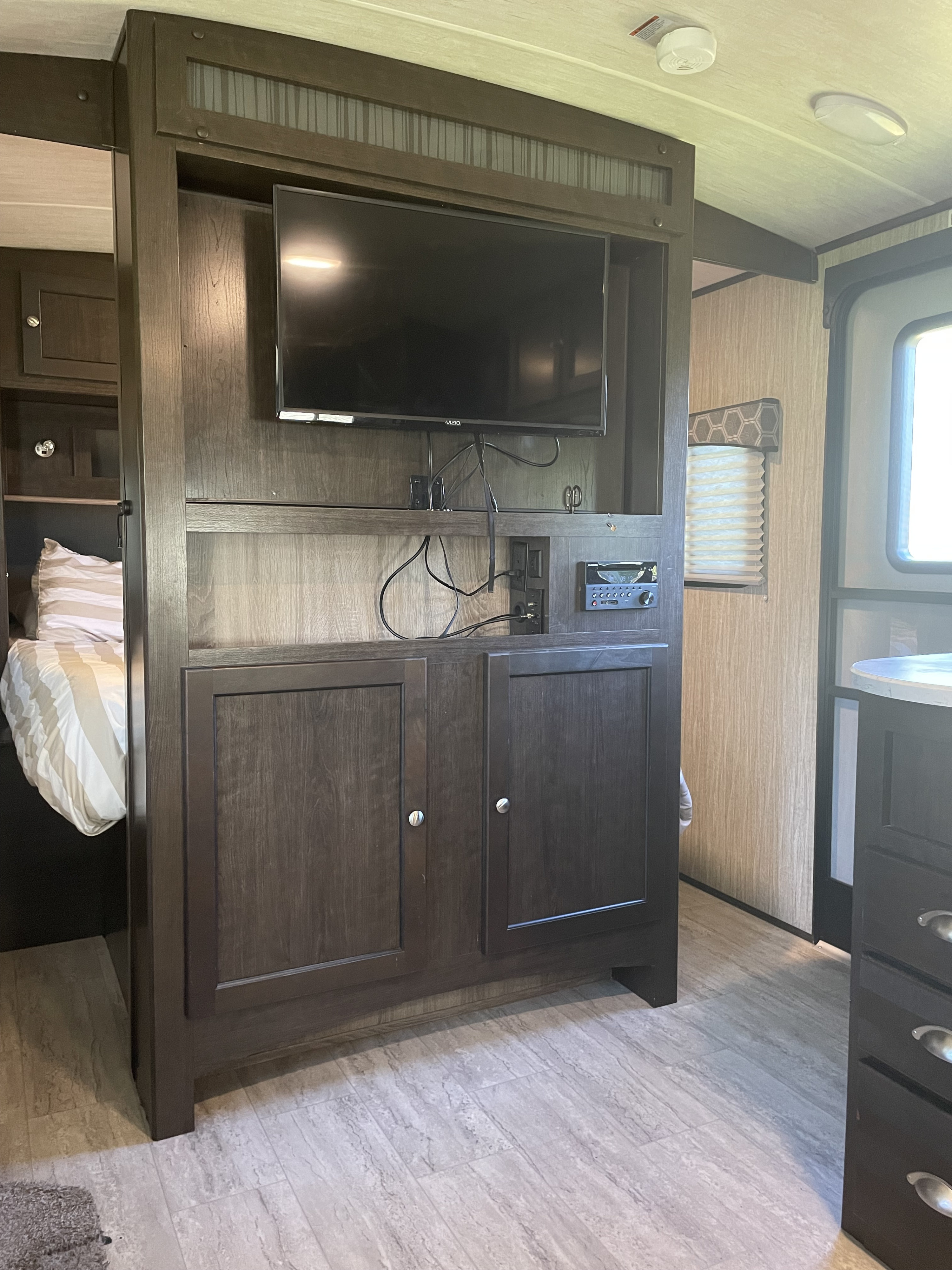 Inside an RV rental: Dark wood entertainment unit with TV, stereo, and cabinets, next to a bed and a bright entryway.. Cruiser RV Shadow Cruiser 2019