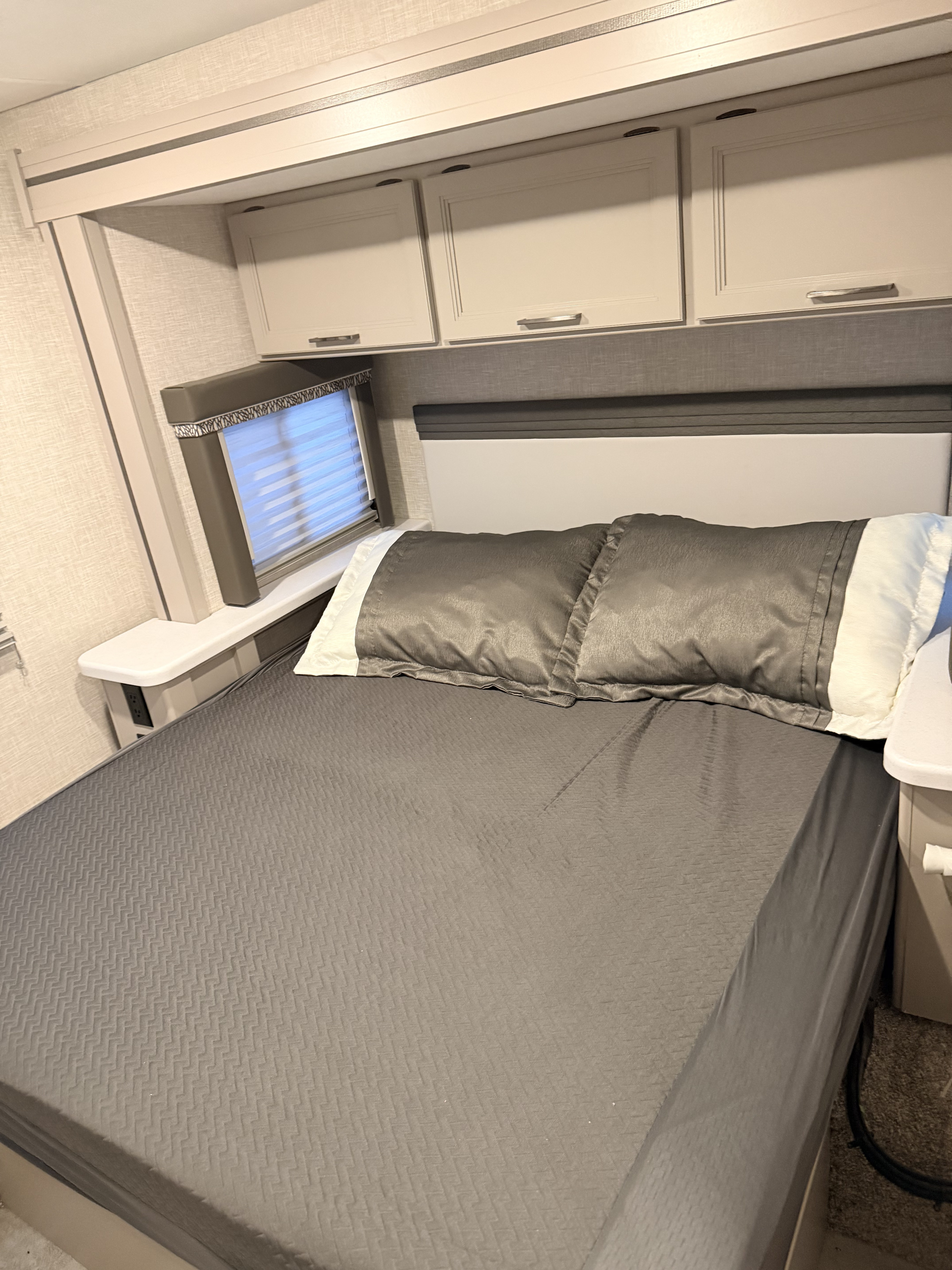 RV rental bedroom with a gray textured bedspread, padded headboard, overhead cabinets, and a window with blinds.. Thor Quantum 2025
