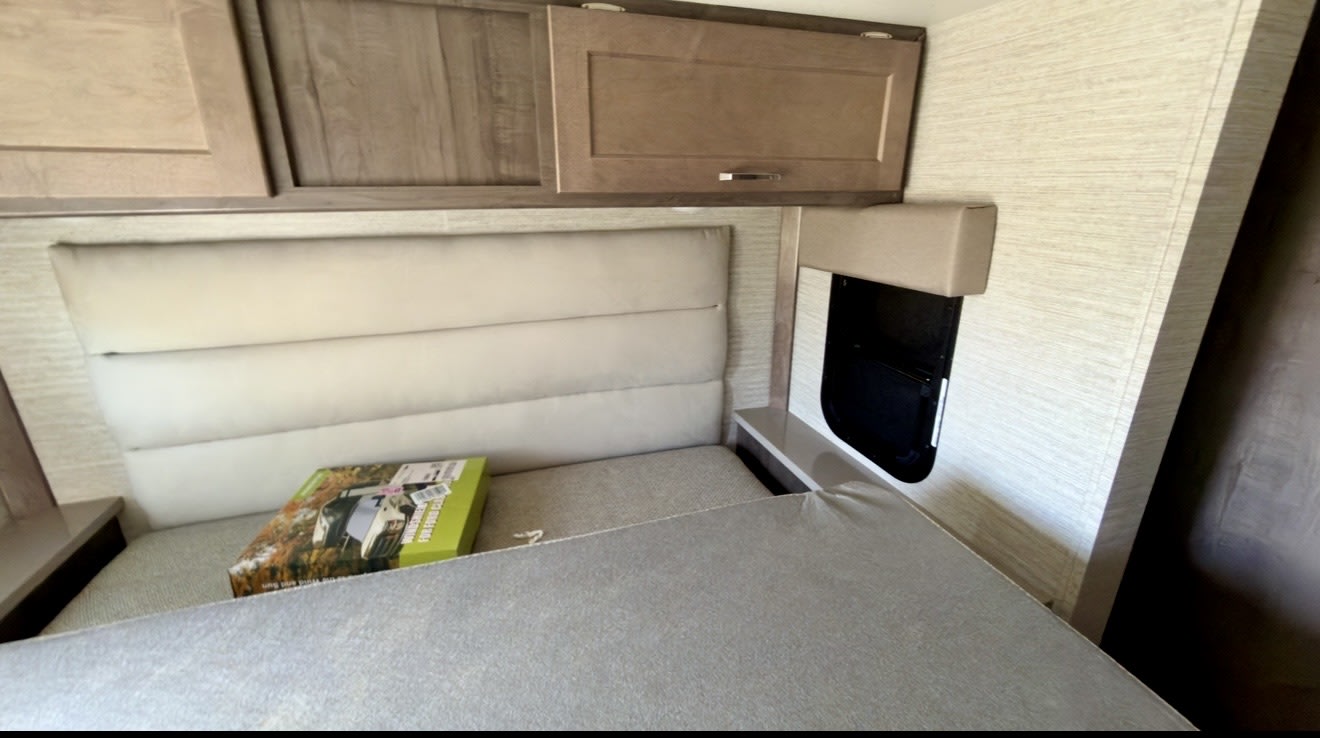 Inside an RV rental: a compact sleeping area with a cream upholstered headboard, wood cabinets, and a small side window. Ready for adventure.. Winnebago Spirit 2021