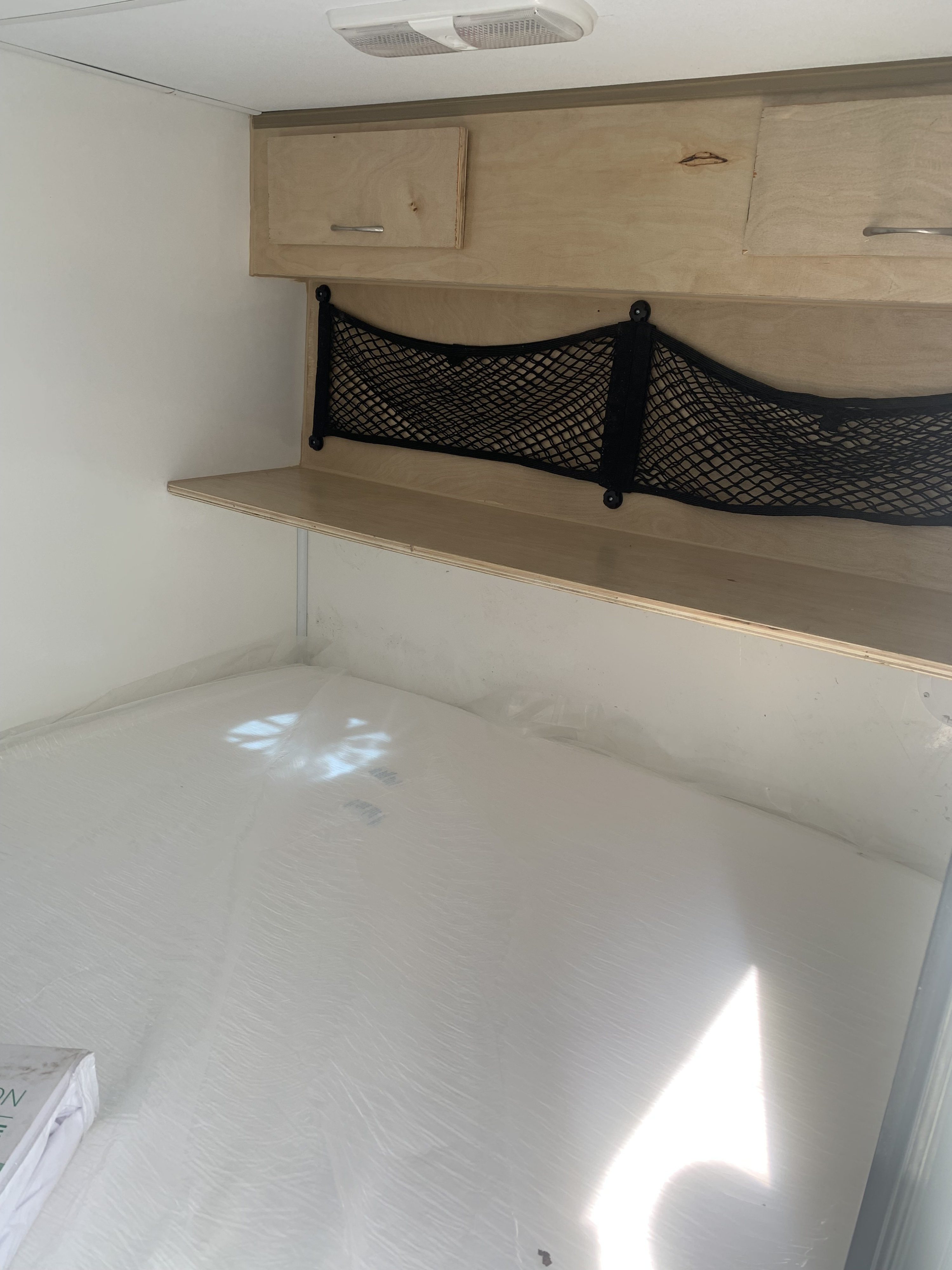 Interior of an RV rental: light wood overhead cabinets, a shelf, black storage net, and a clean white sleeping area.. taroulotte.com teardrop 2026