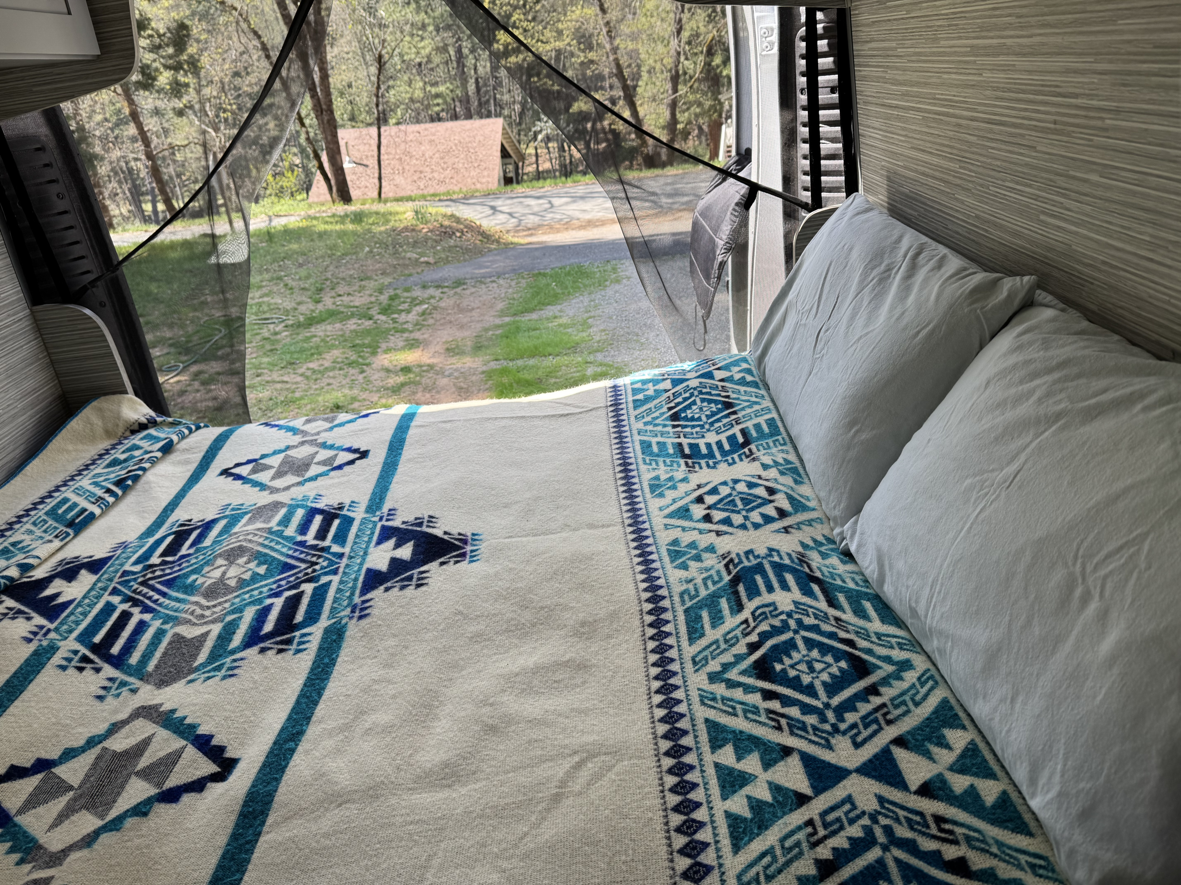Relax in an RV rental! Comfy bed with a blue and white blanket, facing an open screen door with forest views.. Dodge Ram Promaster 2500 High Roof 2019