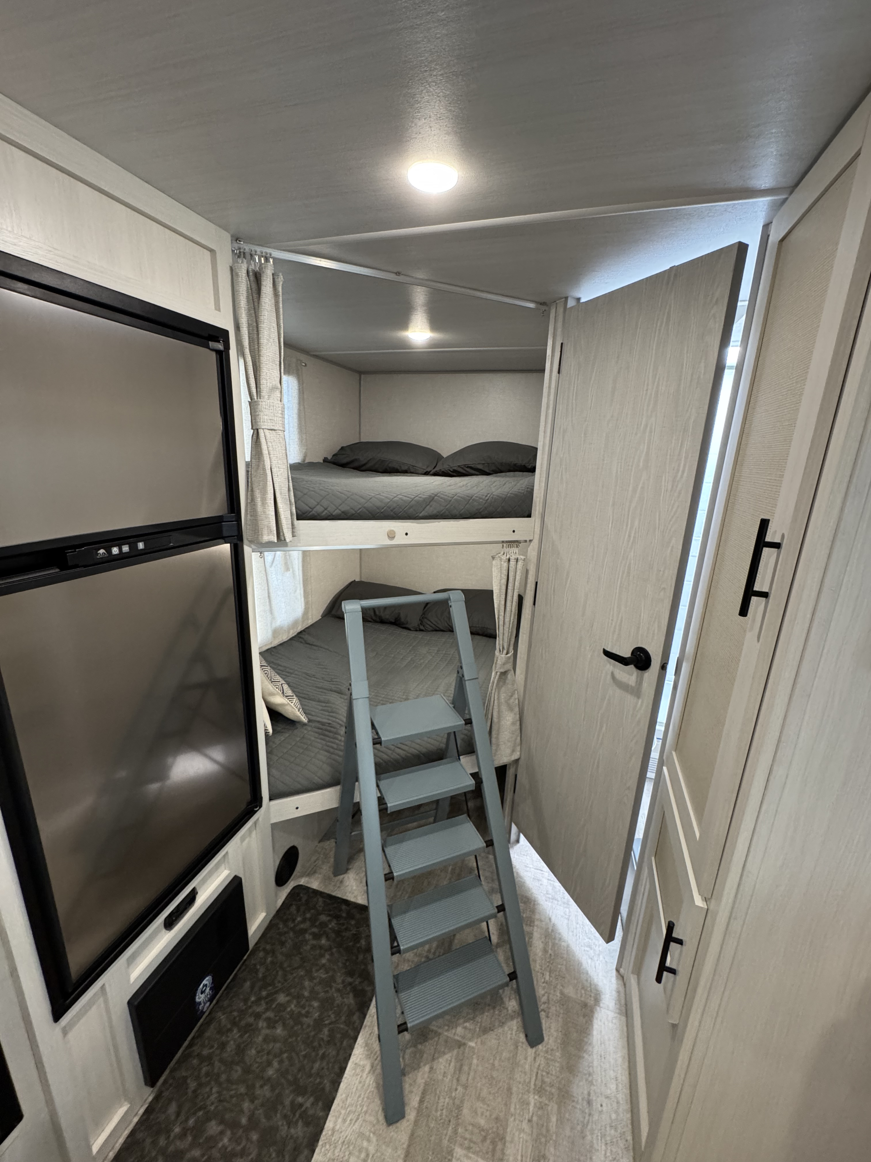 This RV rental bunk area features a modern fridge, two cozy bunks with grey bedding, and a ladder for easy access. Perfect for camping trips.. Forest River No Boundaries Essentials+Only 2024