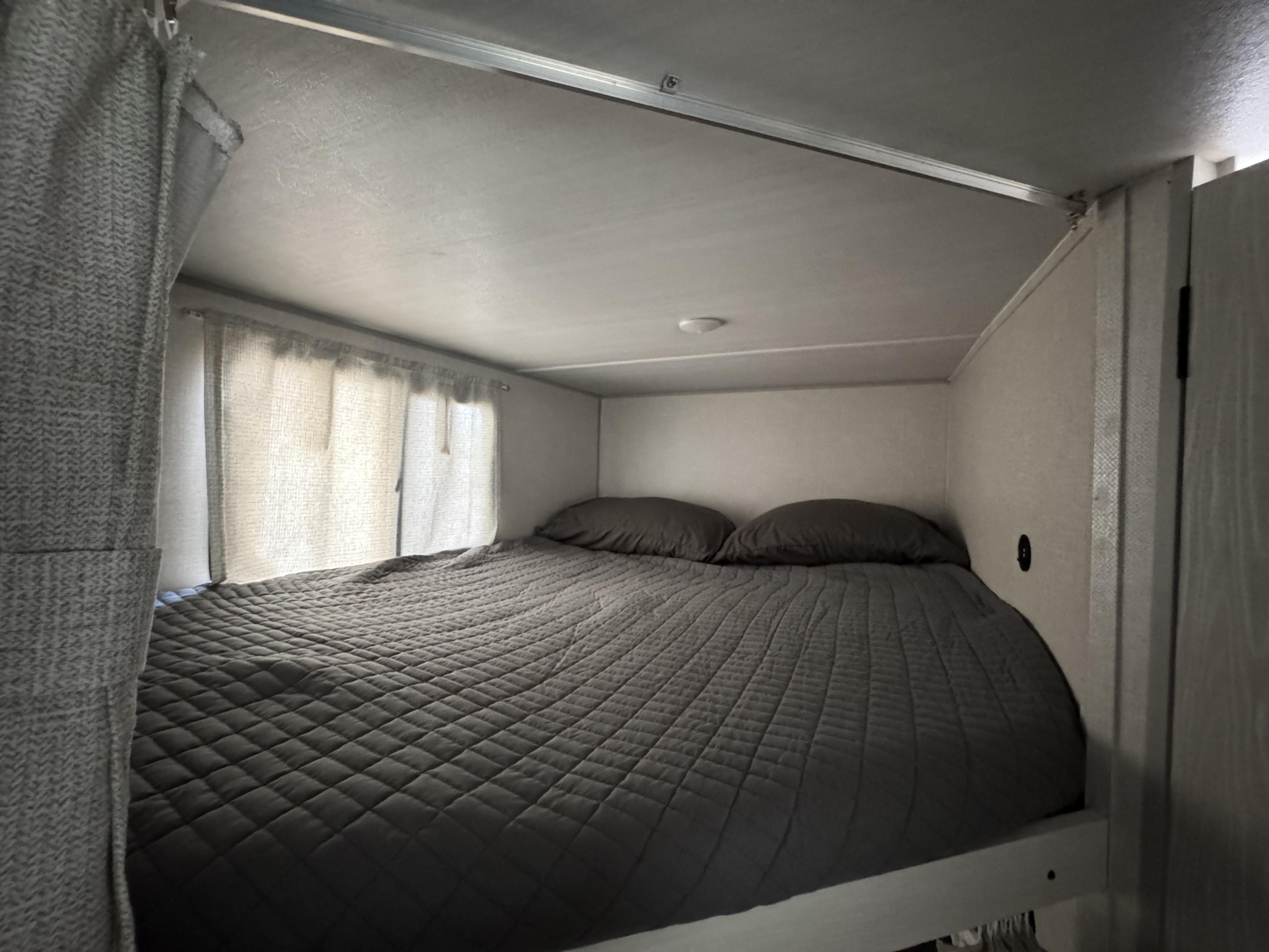 A compact and cozy RV rental bedroom with a quilted bed, pillows, and sheer curtains illuminating the space.. Forest River No Boundaries Essentials+Only 2024