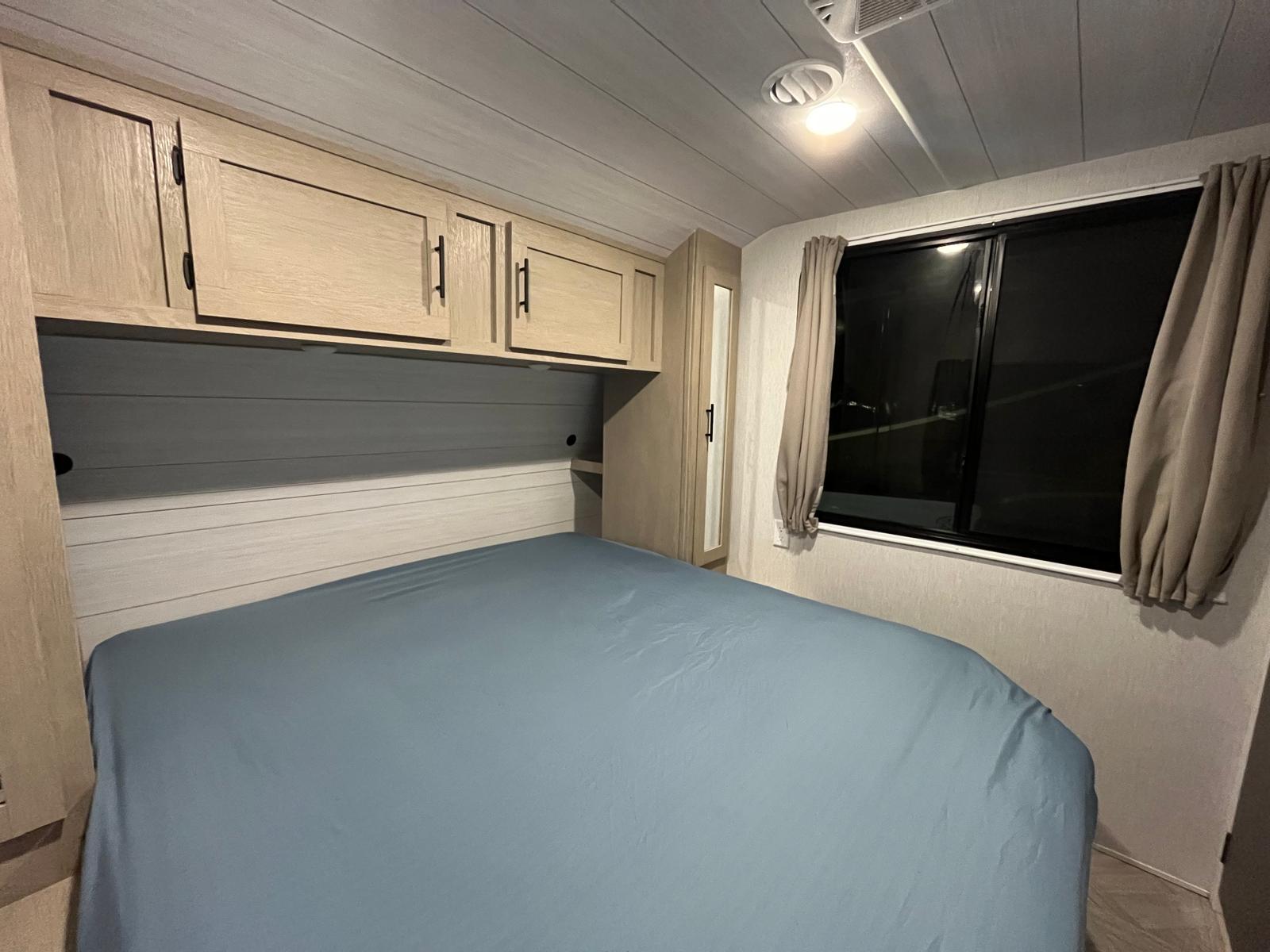 This RV rental bedroom features a blue bed with overhead cabinets, a window with curtains, and mirrored wardrobe storage.. Forest River Wildwood 2024
