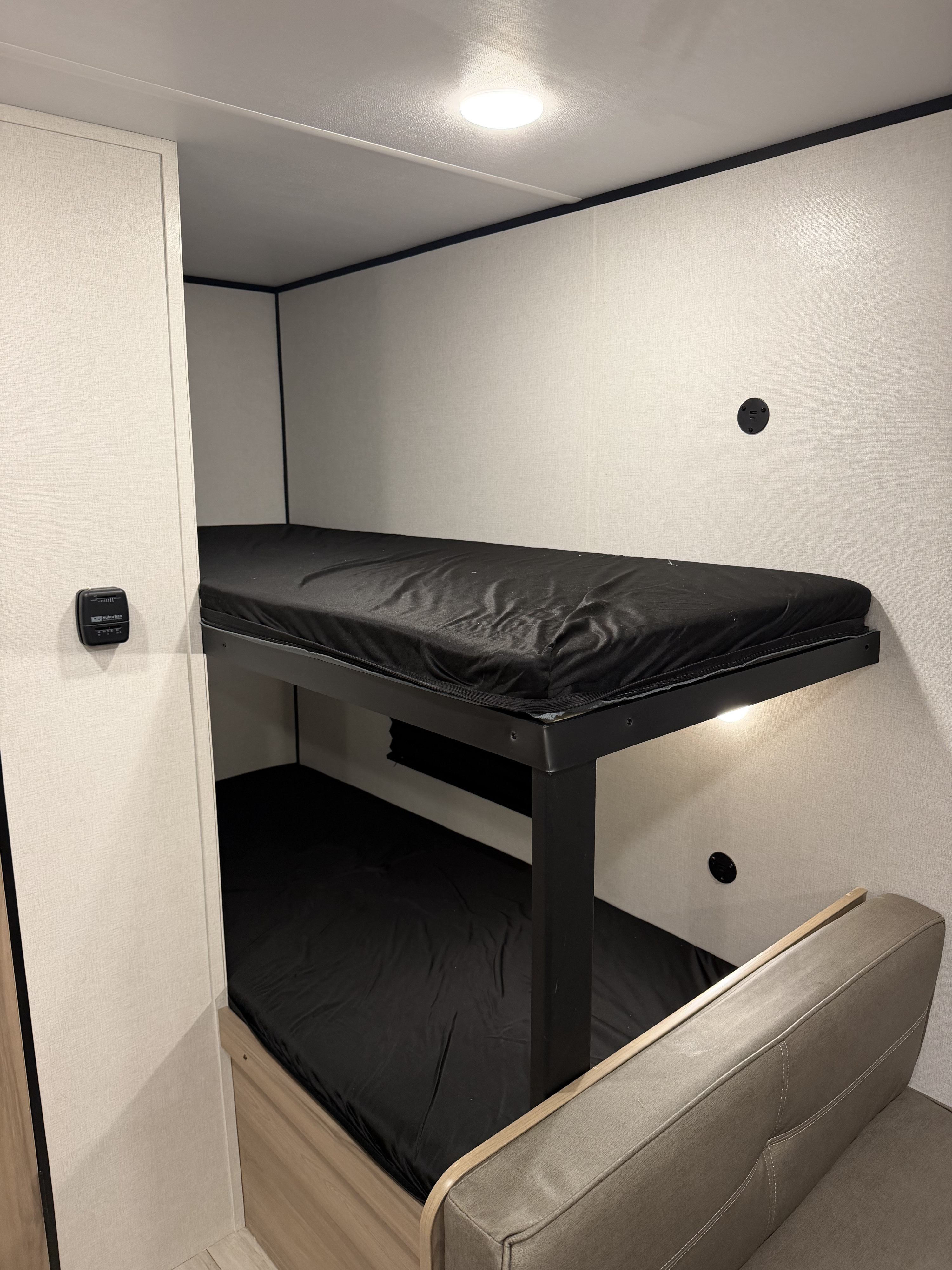 RV rental bunk beds: a compact, functional sleeping area with black mattresses, light walls, a thermostat, and a sofa.. Coleman 17B 2026