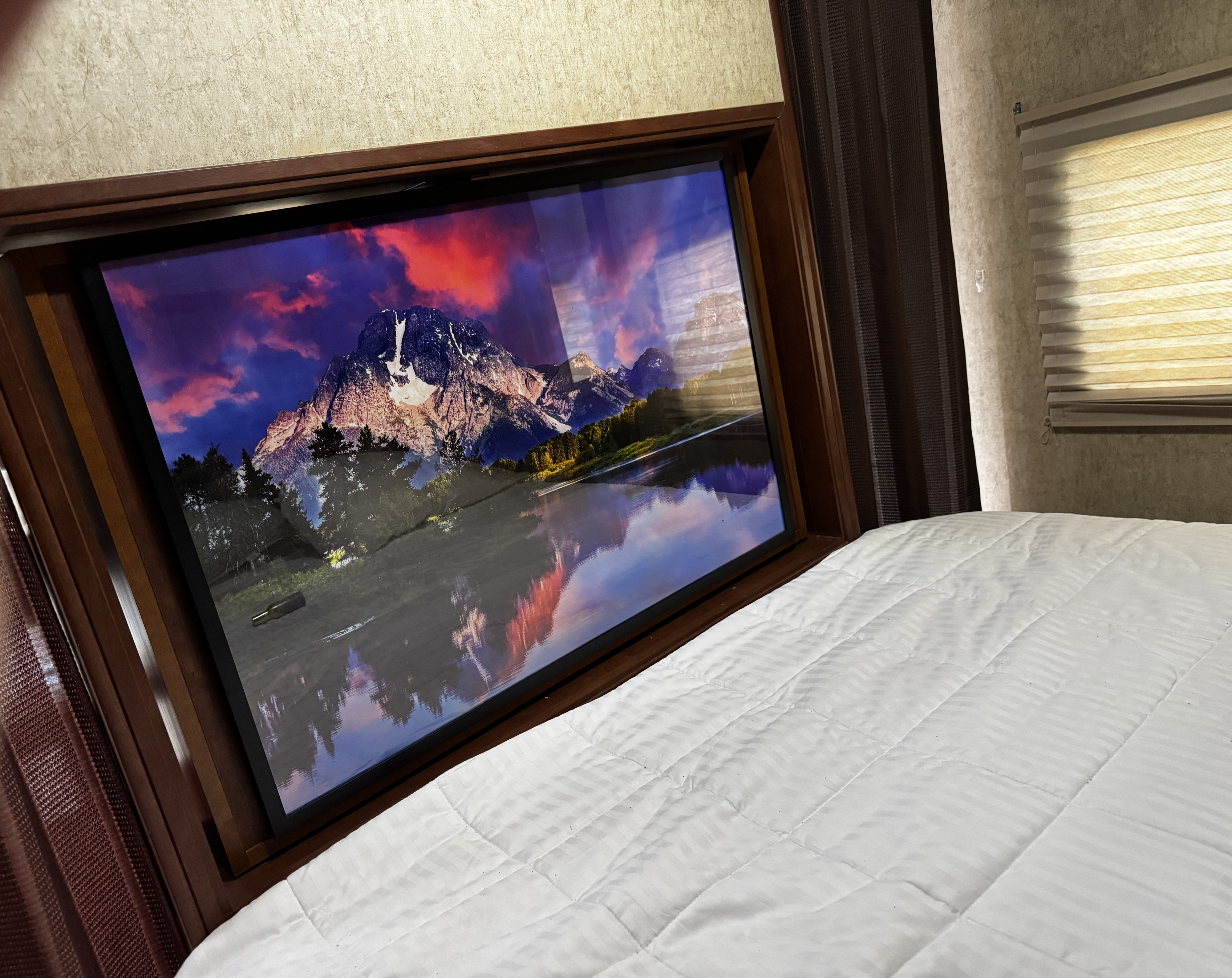 RV rental bedroom: Relax on a comfortable white bed facing a large TV displaying a vibrant mountain landscape with a reflective lake.. Heartland RVs Mallard 2014