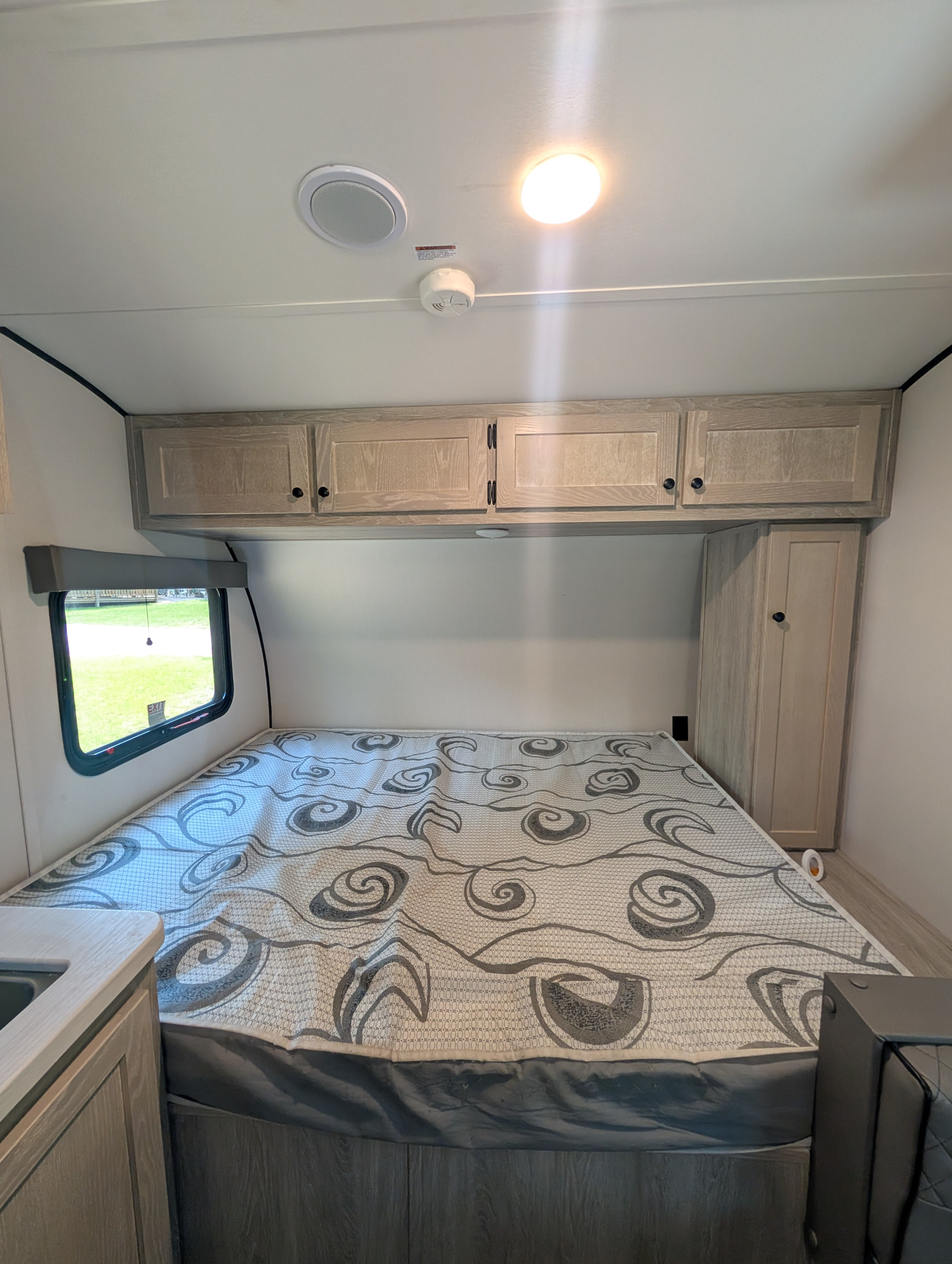 Inside an RV rental: a neat bedroom with a patterned mattress, wooden overhead cabinets, and a side window.. Riverside RV Xplorer 2024