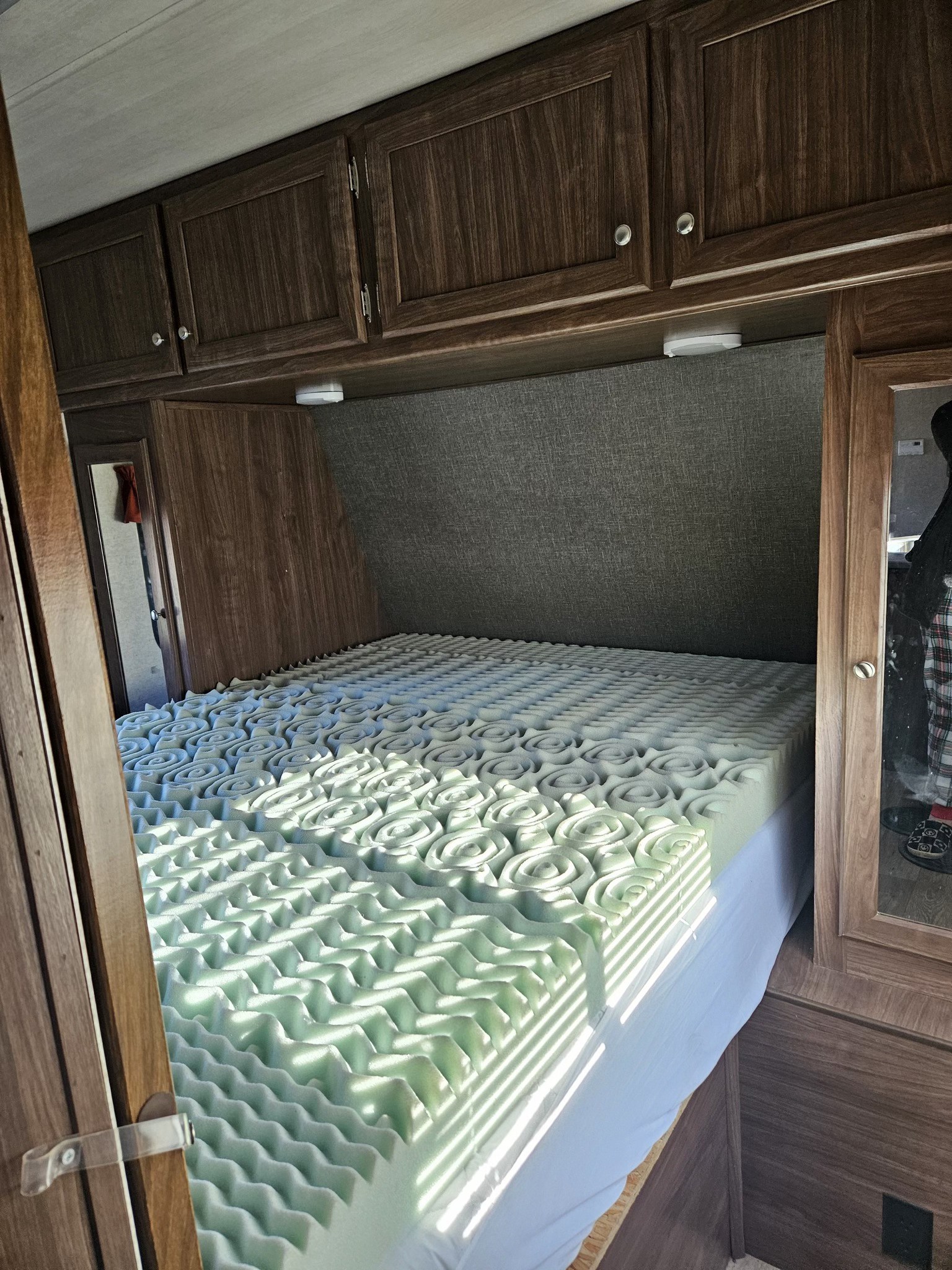 Inside a comfortable RV rental bedroom, featuring a large bed with a light green textured foam topper and overhead wooden cabinets.. Heartland RVs Prowler 2018