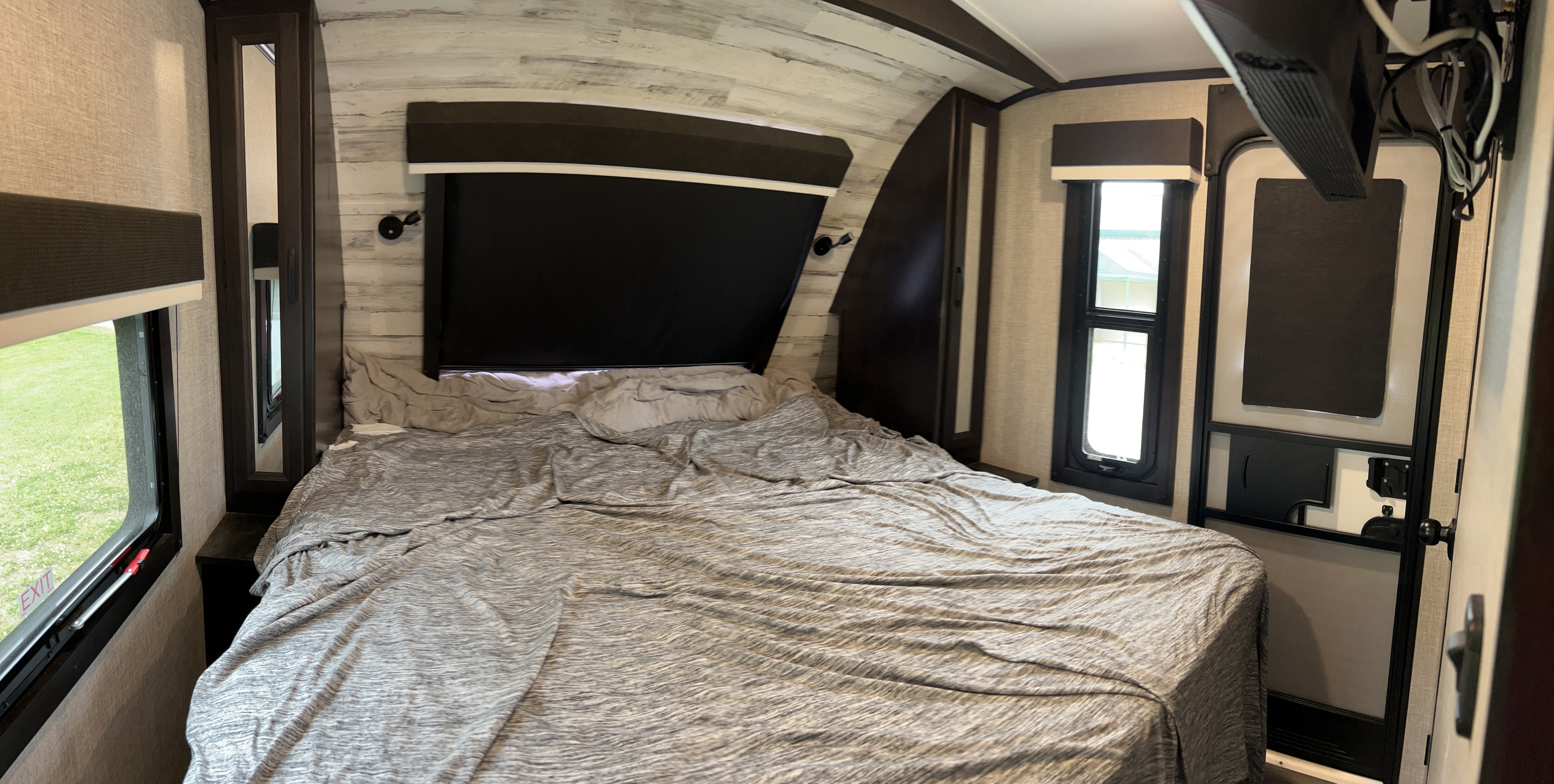 This RV rental bedroom features a large bed with a grey comforter, windows, a dark headboard, and a mounted TV.. Jayco White Hawk 2022