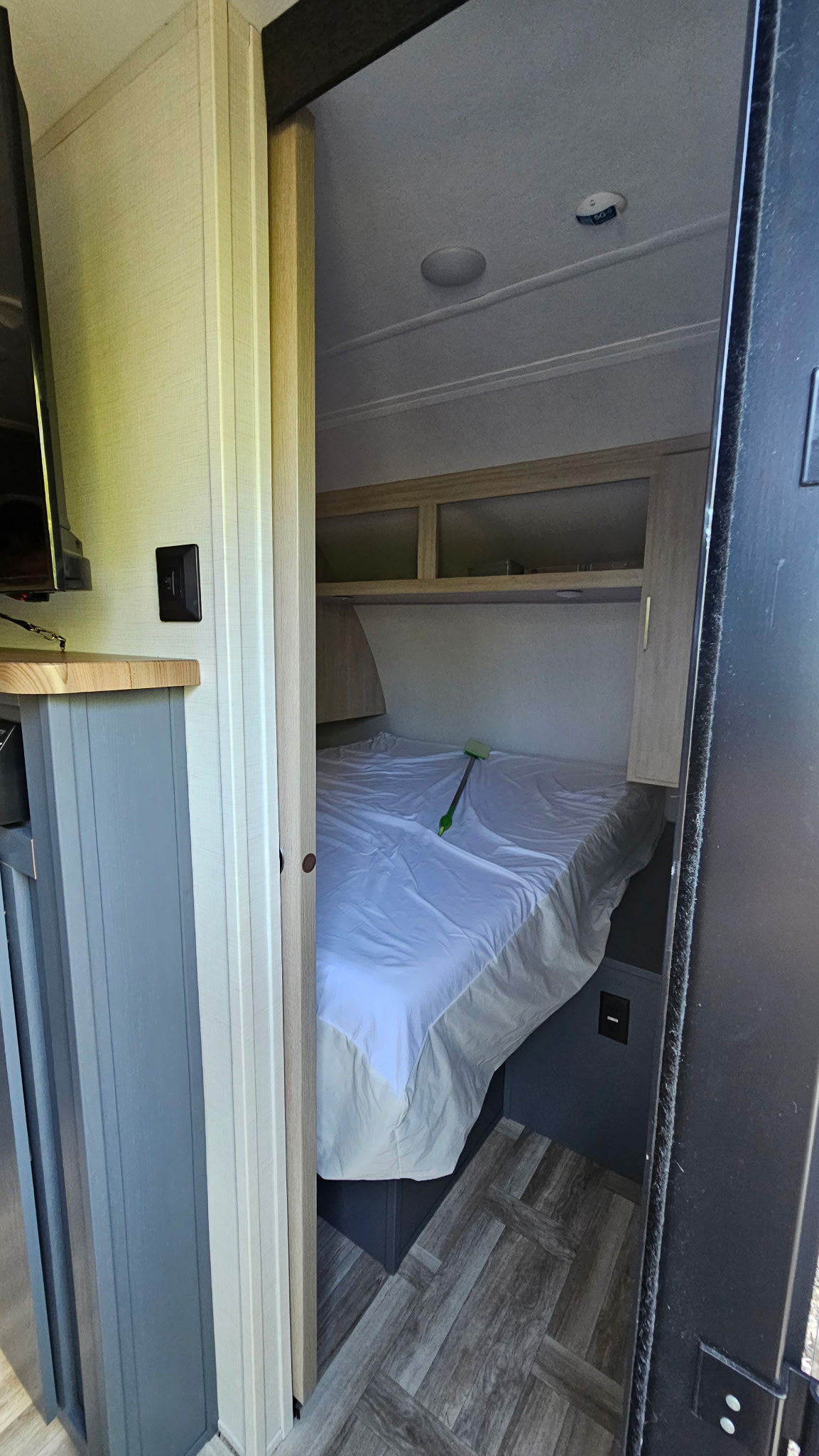 View into an RV rental bedroom: white-sheeted bed, overhead cabinets, ceiling light, smoke detector, and wood-look floor.. Forest River Tracer 2024