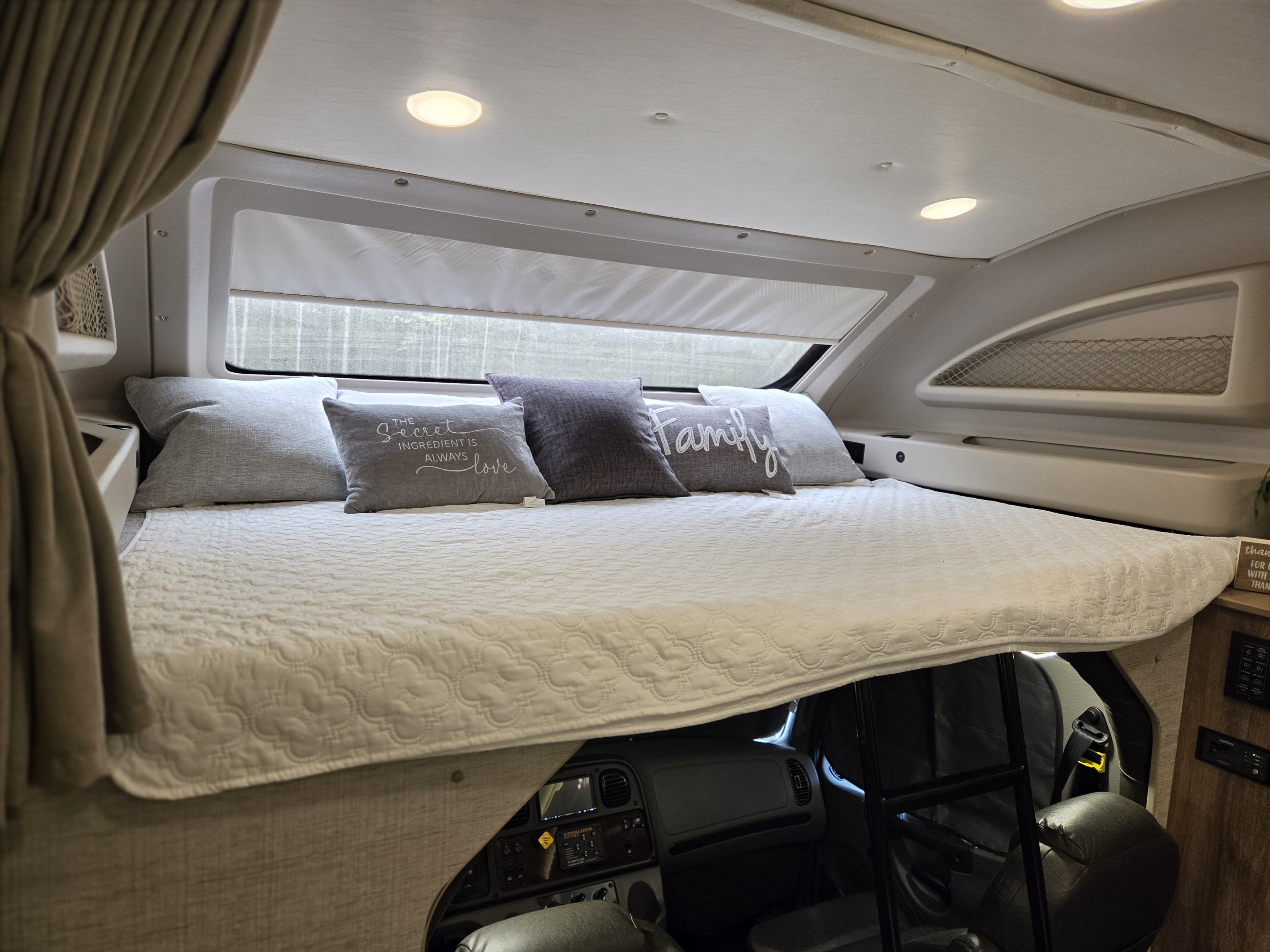 Cozy over-cab bunk bed in an RV rental, with decorative pillows, a window view, and the cockpit visible below.. Jayco Seneca 2021