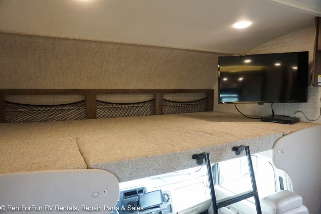Inside an RV rental: a comfortable over-cab bunk bed with TV, storage nets, and access ladder. Ideal for travel.. Jayco Redhawk 2021