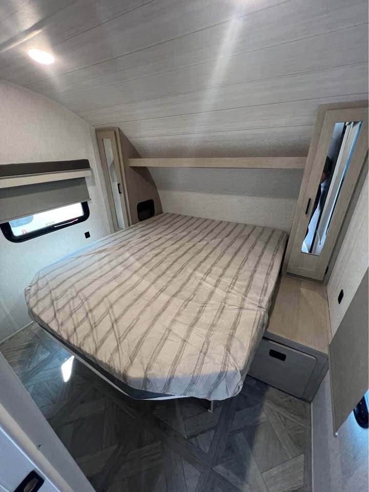 Bright RV rental bedroom with a striped queen bed, mirrored storage, window, and dark patterned floor.. Forest River Wildwood X-Lite 2023