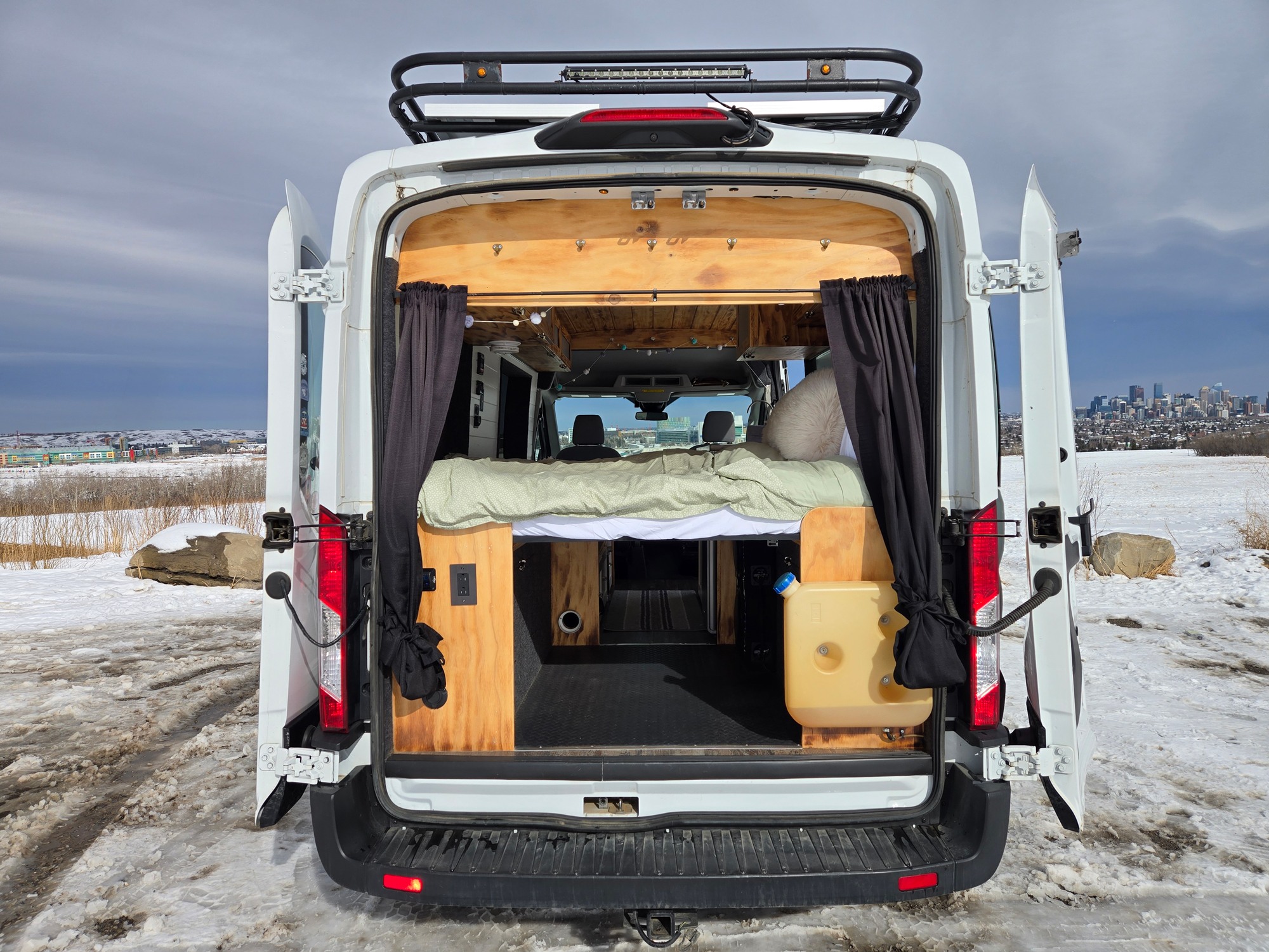 Open rear doors of an RV rental camper van reveal a cozy, wooden interior with a bed, parked in a snowy landscape with a distant city skyline.. Ford Transit T-250 2019