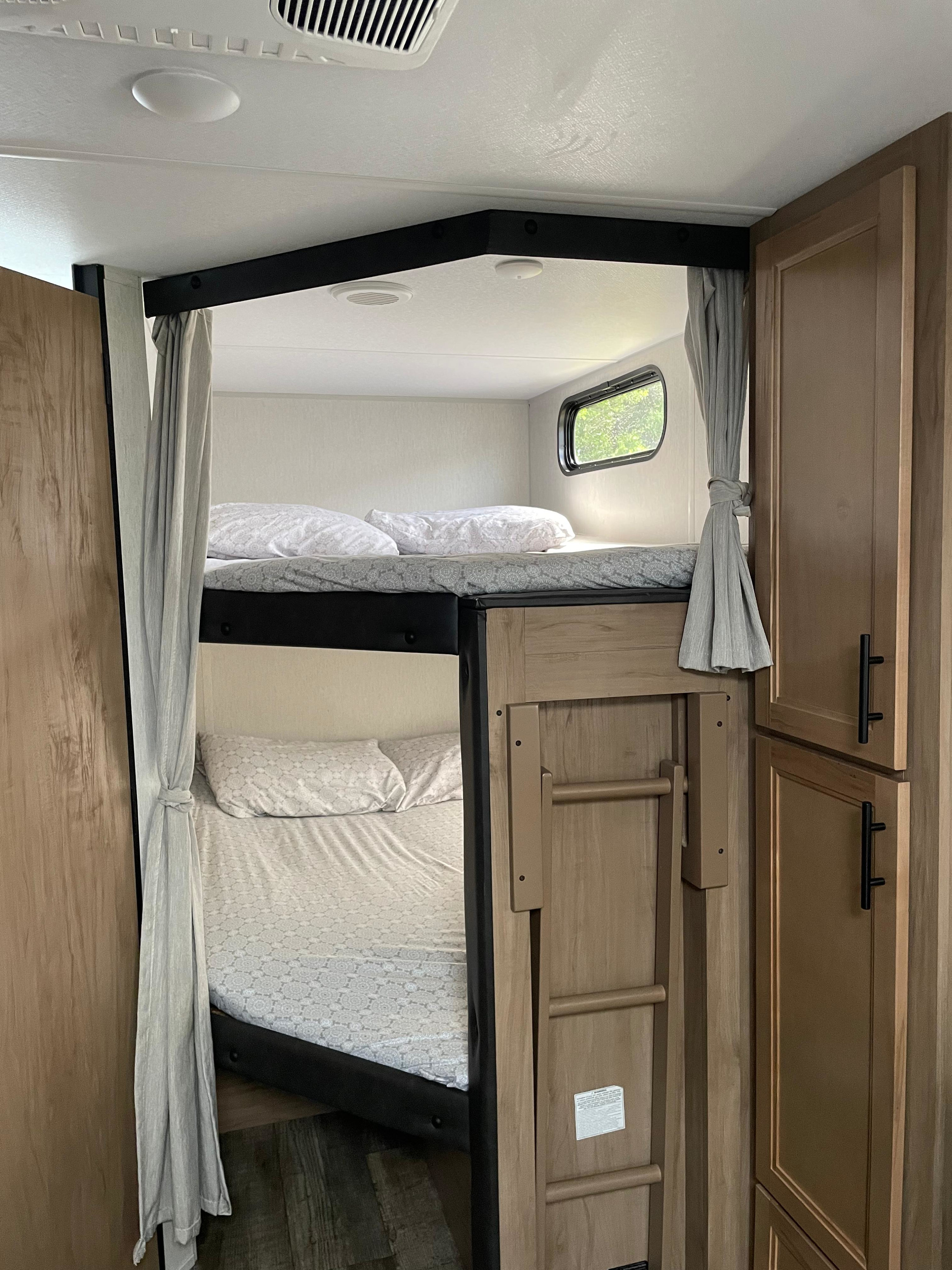 Inside an RV rental, showing neat bunk beds with soft bedding, privacy curtains, a window, and a ladder next to wooden cabinets.. Forest River Cherokee Grey Wolf 2023