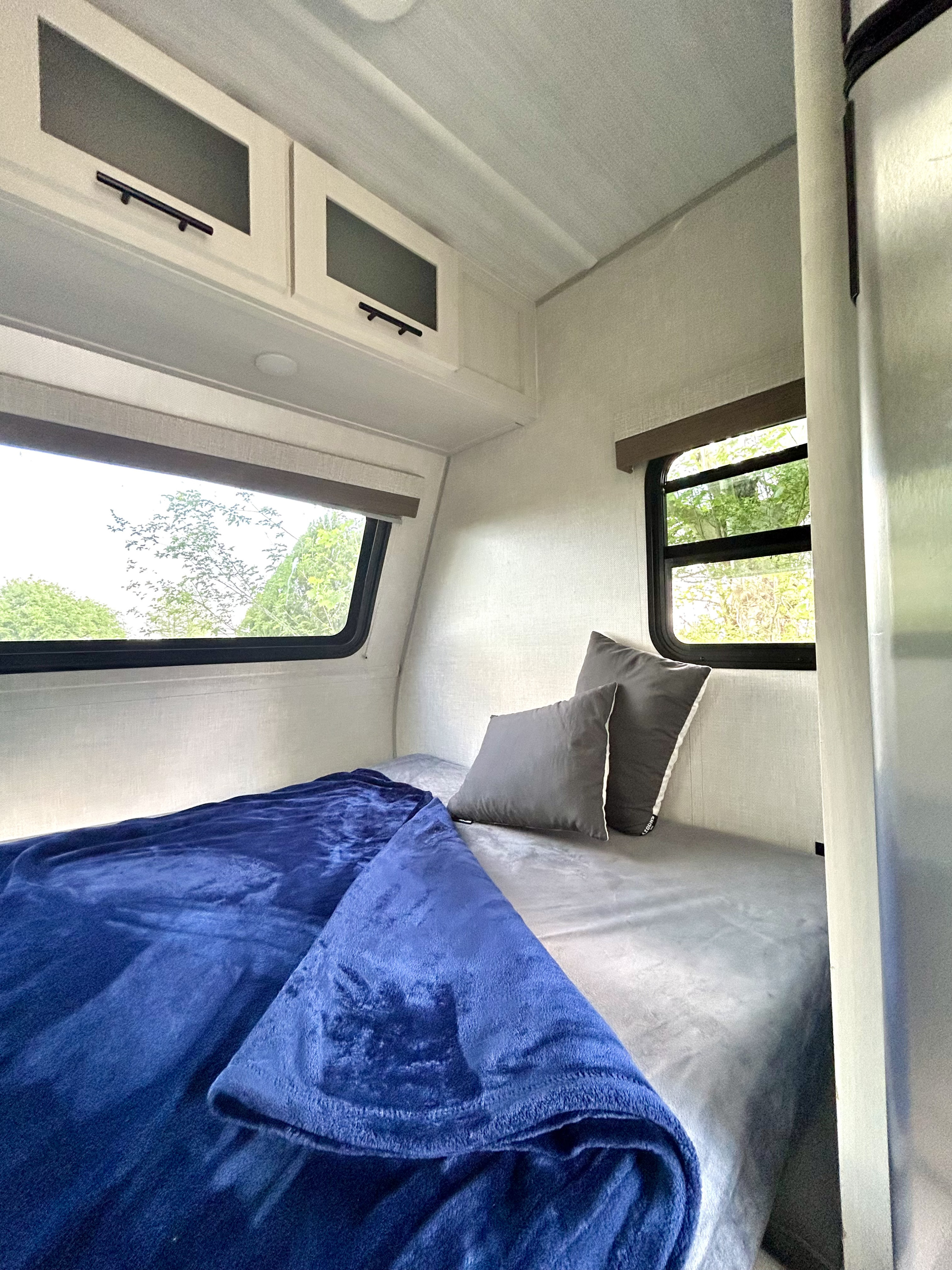 Enjoy this RV rental's cozy bedroom: a bed with a soft blue blanket, gray pillows, and sunny windows overlooking trees.. Forest River No Boundaries 16.1 2023
