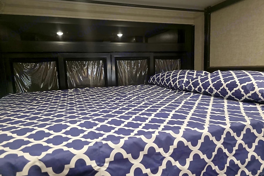 This RV rental bedroom features a comfy bed with blue geometric bedding and a dark headboard with protective covers.. Heartland RVs Cyclone HD Edition Toy Hauler 2015