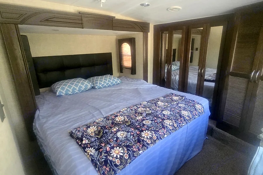 Comfortable RV rental bedroom: large bed with blue bedding, floral blanket, dark headboard, mirrored closets. Cozy interior.. Heartland RVs Cyclone HD Edition Toy Hauler 2015