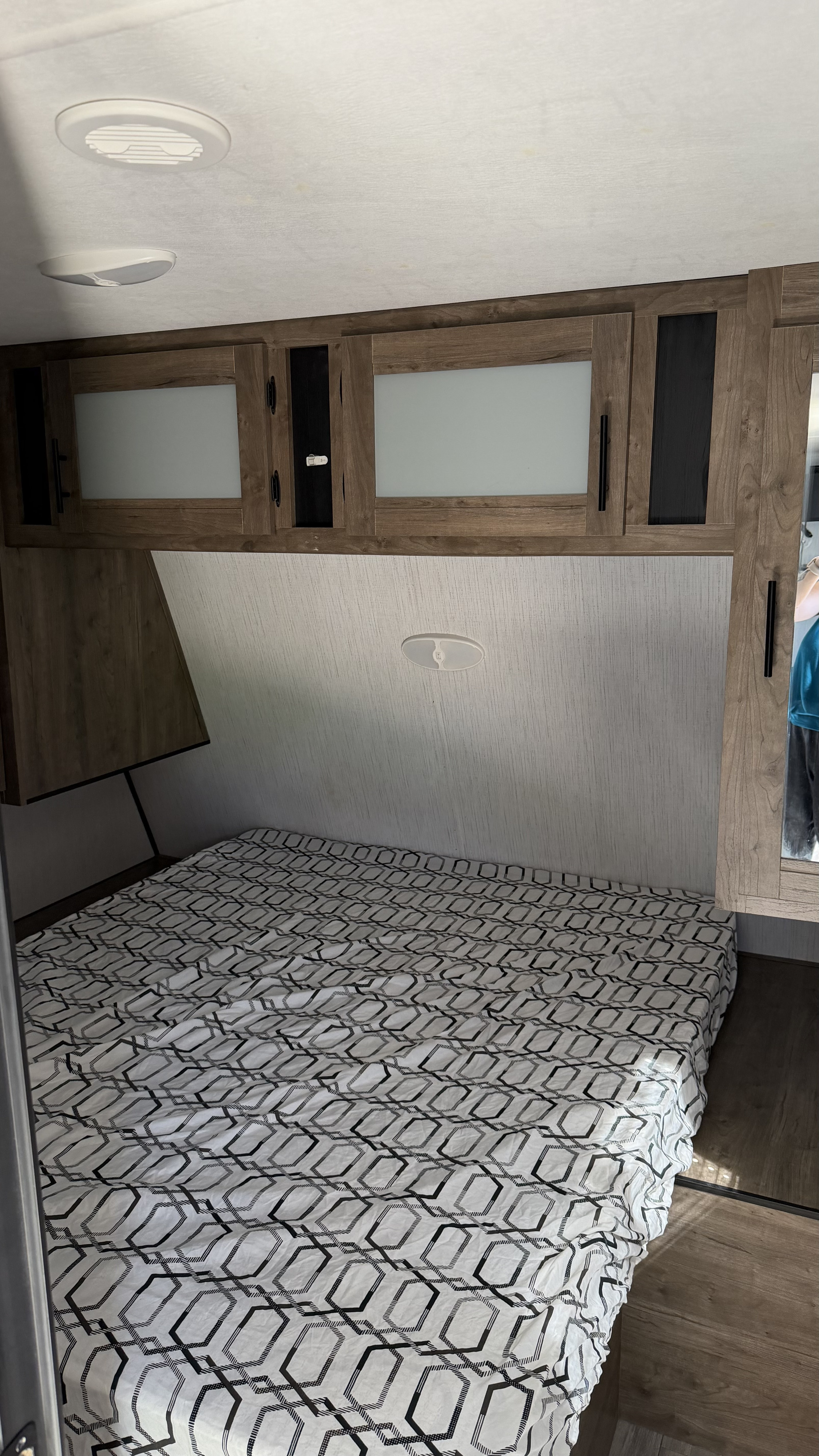 RV rental bedroom with a white and black geometric bedspread, wooden overhead cabinets, and ceiling lights.. Heartland RVs Pioneer 2021