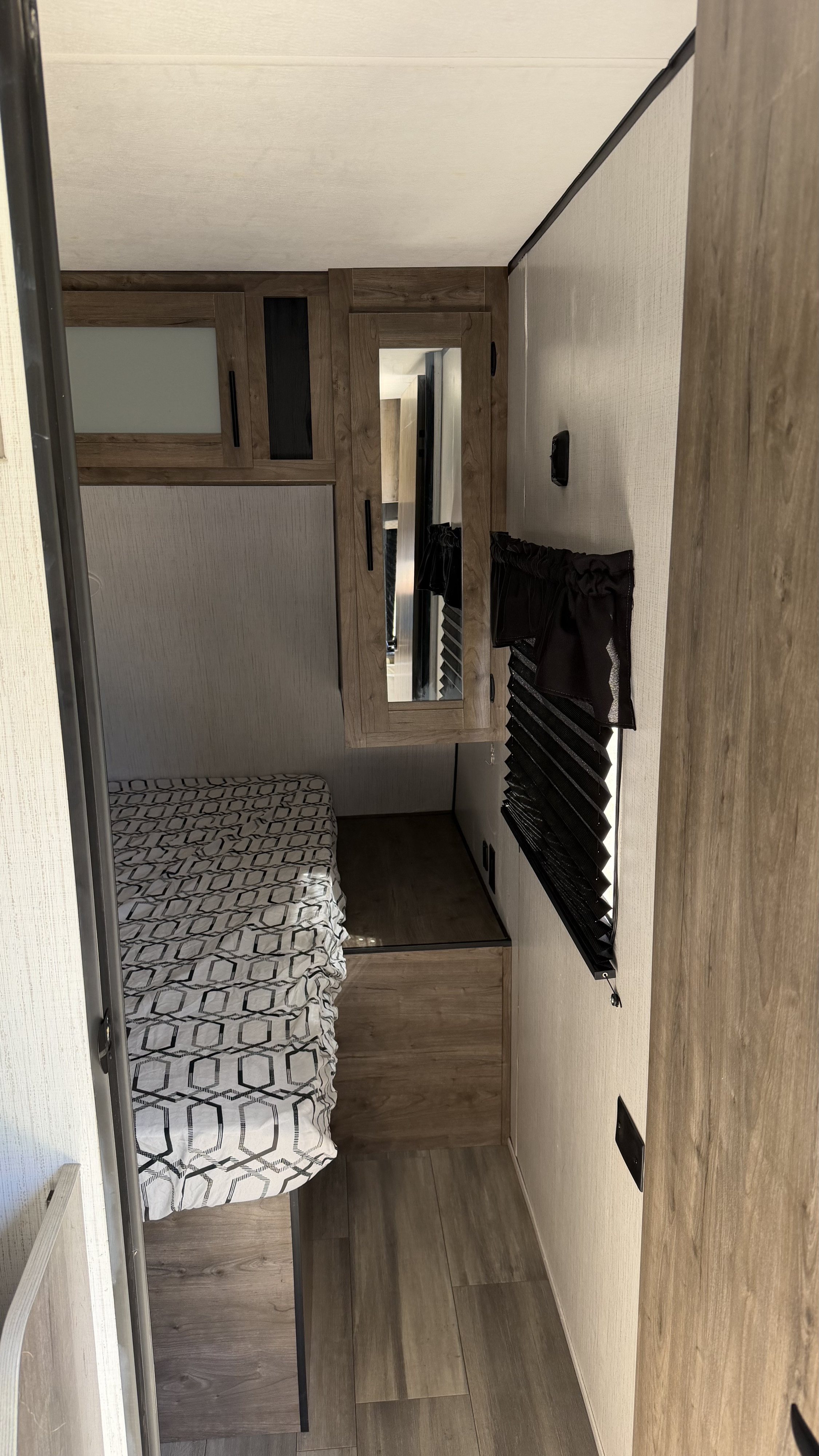 RV rental bedroom featuring a patterned bed, wood cabinetry with a mirror, window with blind, and wood-look floor.. Heartland RVs Pioneer 2021