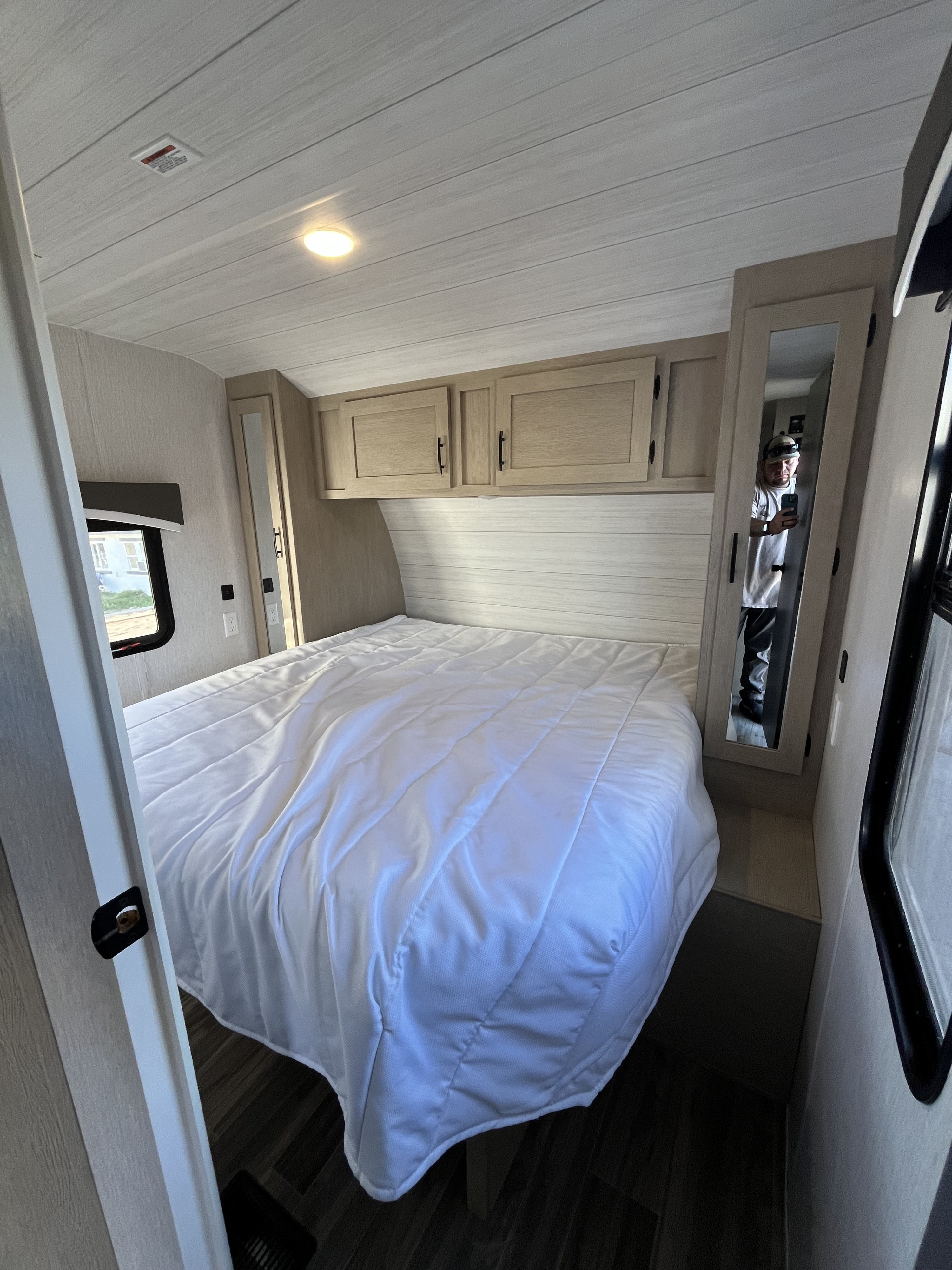 Cozy RV rental bedroom with white bed, overhead storage, and light-colored interior. A person is reflected in the mirror.. Forest River Evo 2700 2023