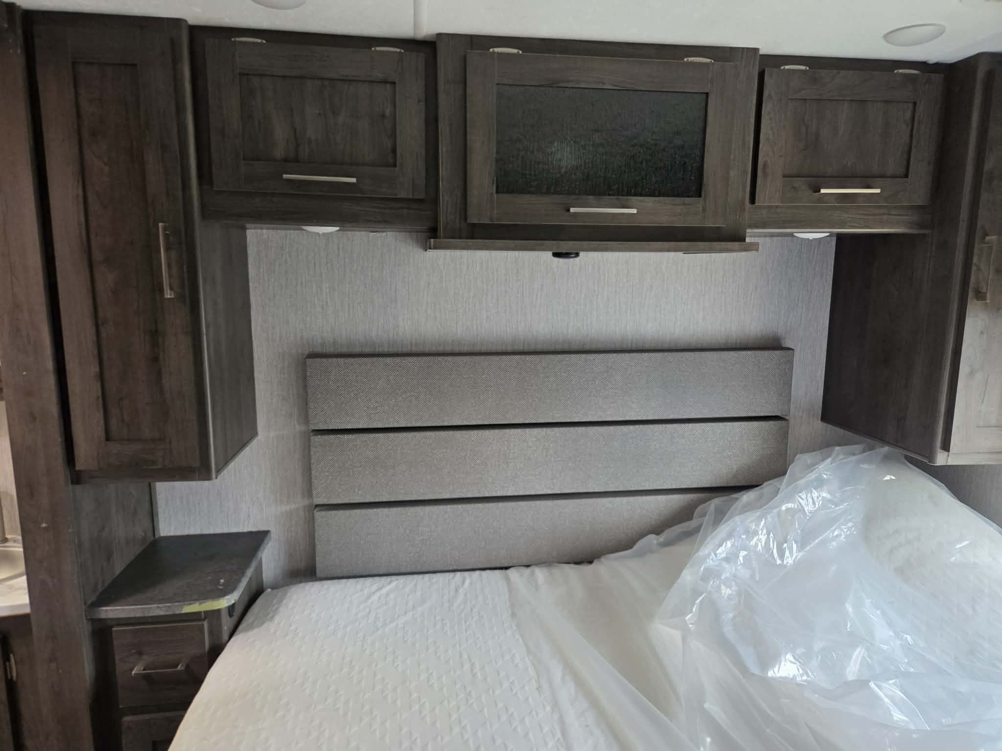 Inside an RV rental: a cozy bedroom with dark wood overhead cabinets, a textured headboard, and a new mattress.. Forest River Coachmen Pursuit 2022