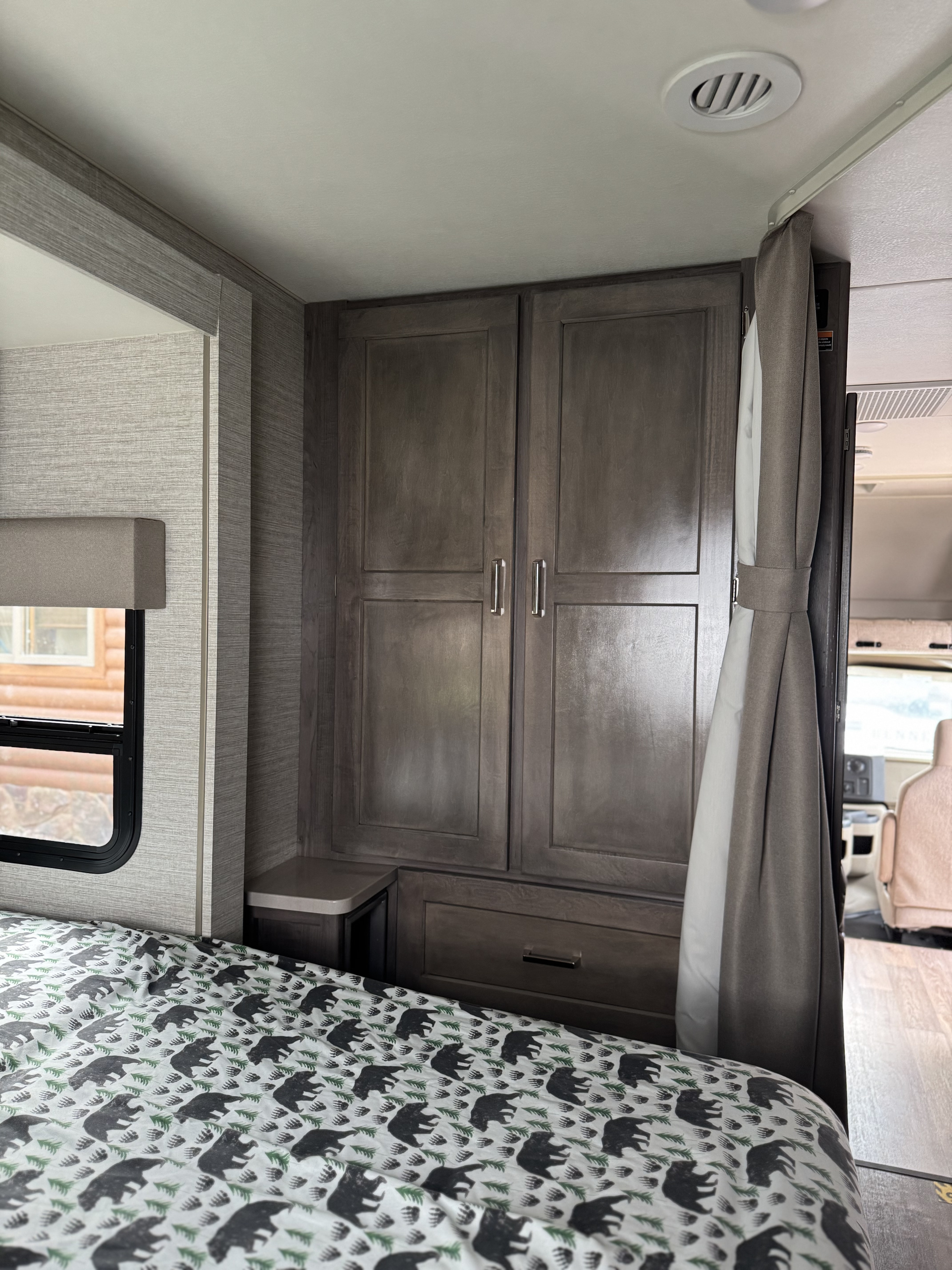 RV rental bedroom featuring a bed with bear-patterned duvet, dark wood wardrobe storage, and a side window.. Winnebago Minnie Winnie 2020
