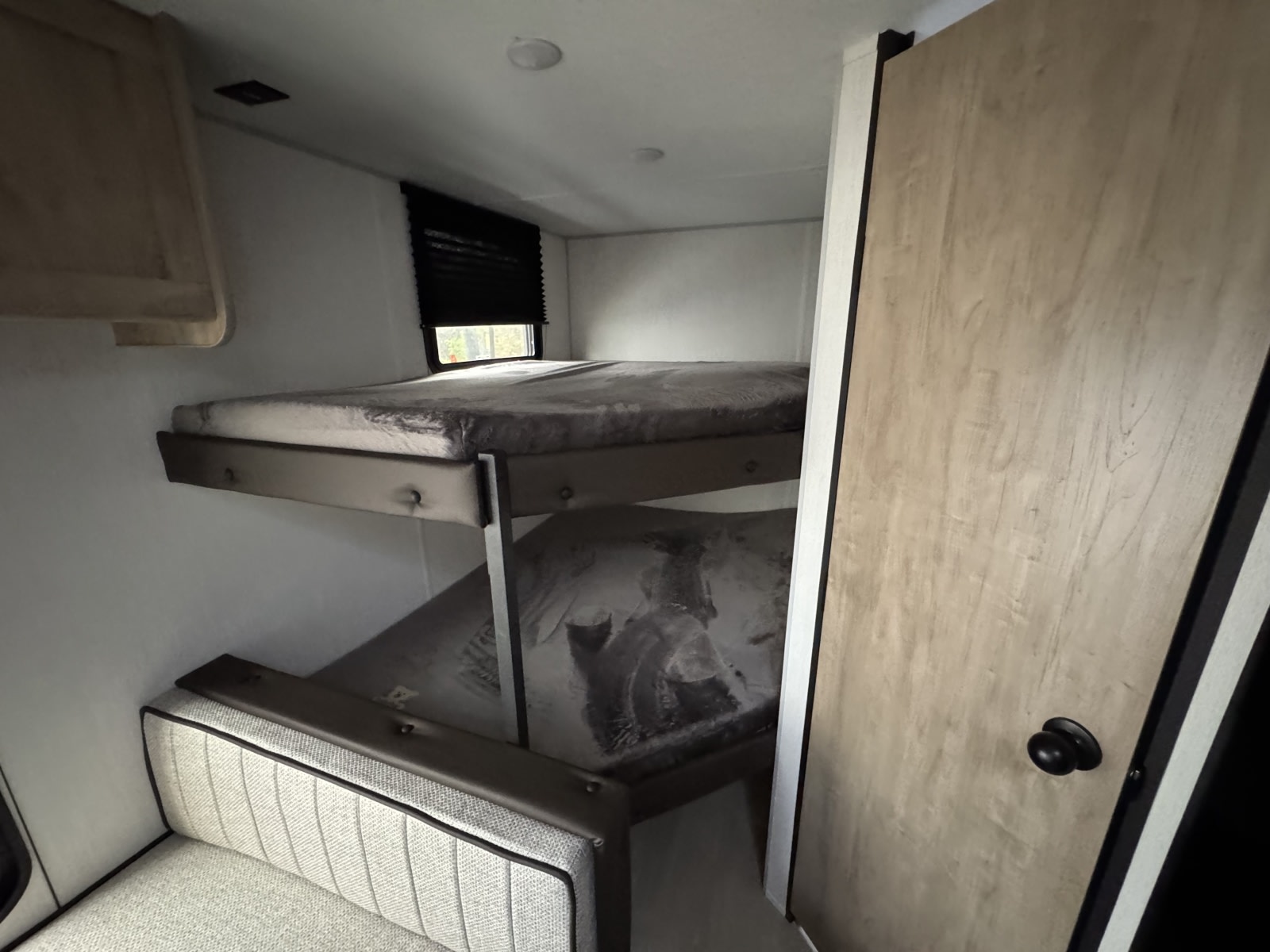 RV rental interior: compact sleeping area with bunk beds, a window with blinds, and an upholstered bench seat by a wooden door.. Forest River GREY WOLF 26LP 2026