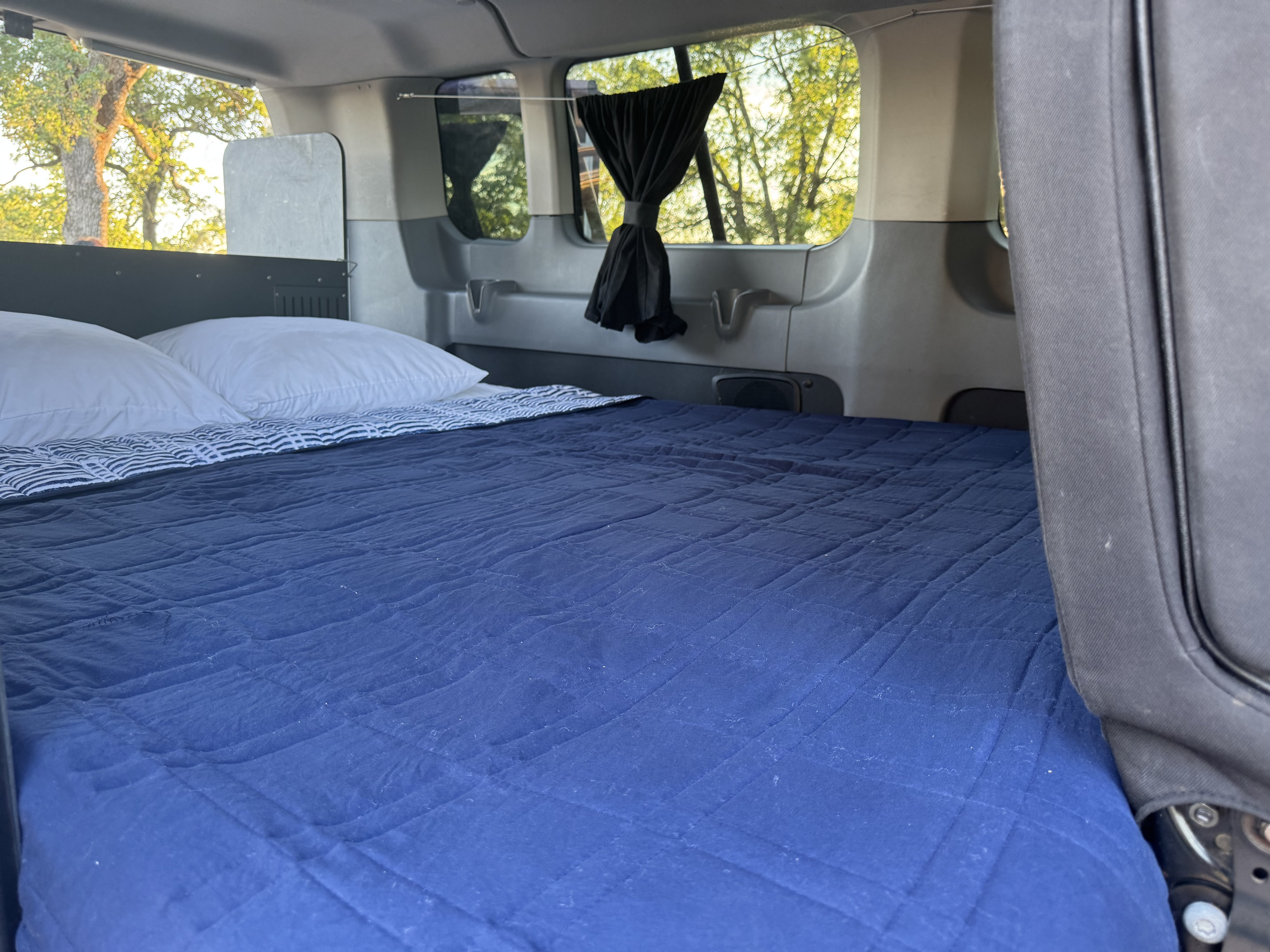 Inside an RV rental: a comfy bed with blue quilted bedding and white pillows. Windows with curtains offer tree views.. Ford Transit custom 2017