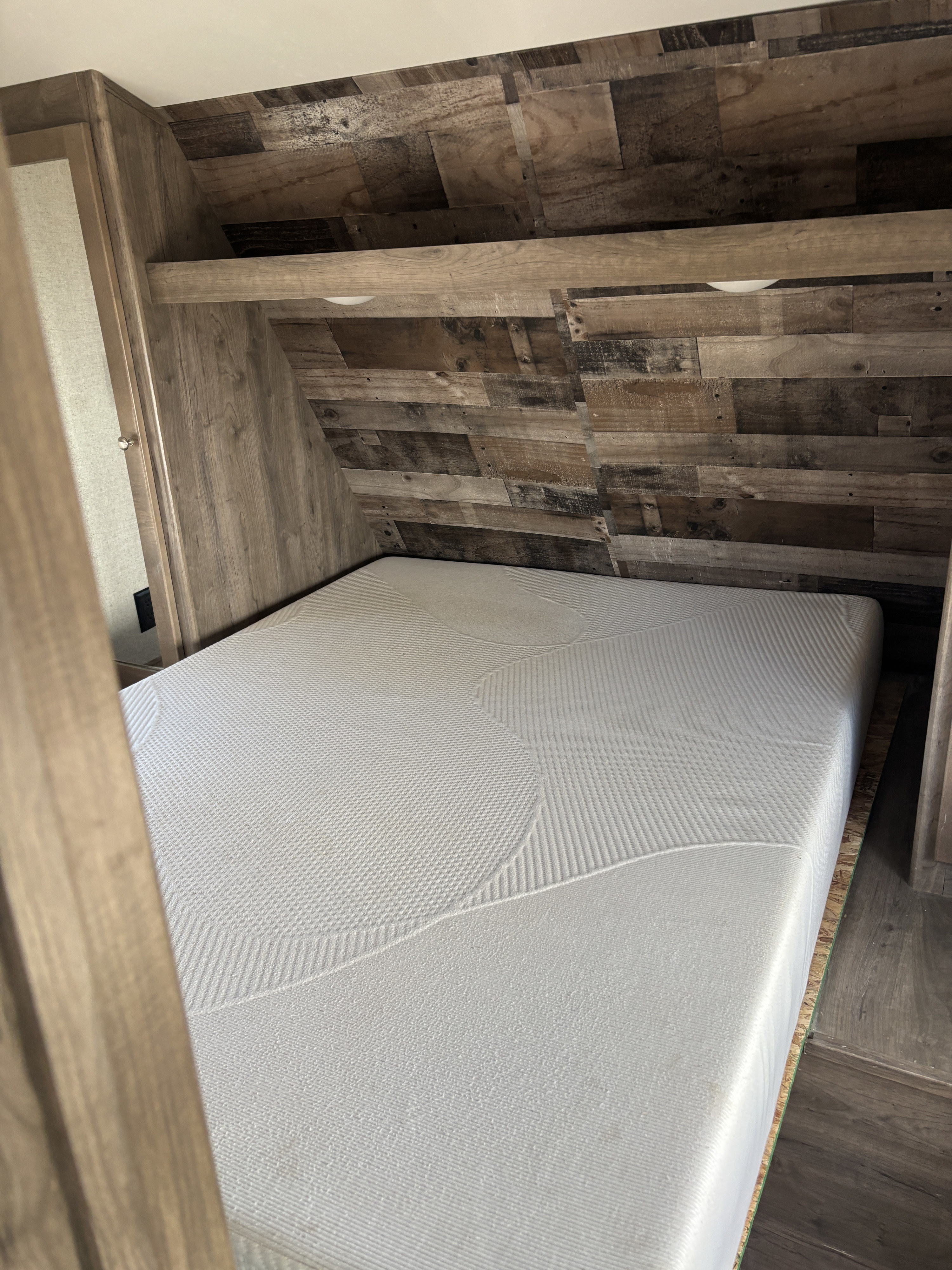 Inside an RV rental, a comfy white mattress on a bed base, surrounded by rustic wooden walls and a shelf with lights.. Dutchmen Aspen Trail 2019