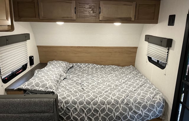 Experience comfort in this RV rental bedroom, featuring a cozy bed, wooden headboard, overhead cabinets, and two windows.. Forest River Cherokee Wolf Pup 2024
