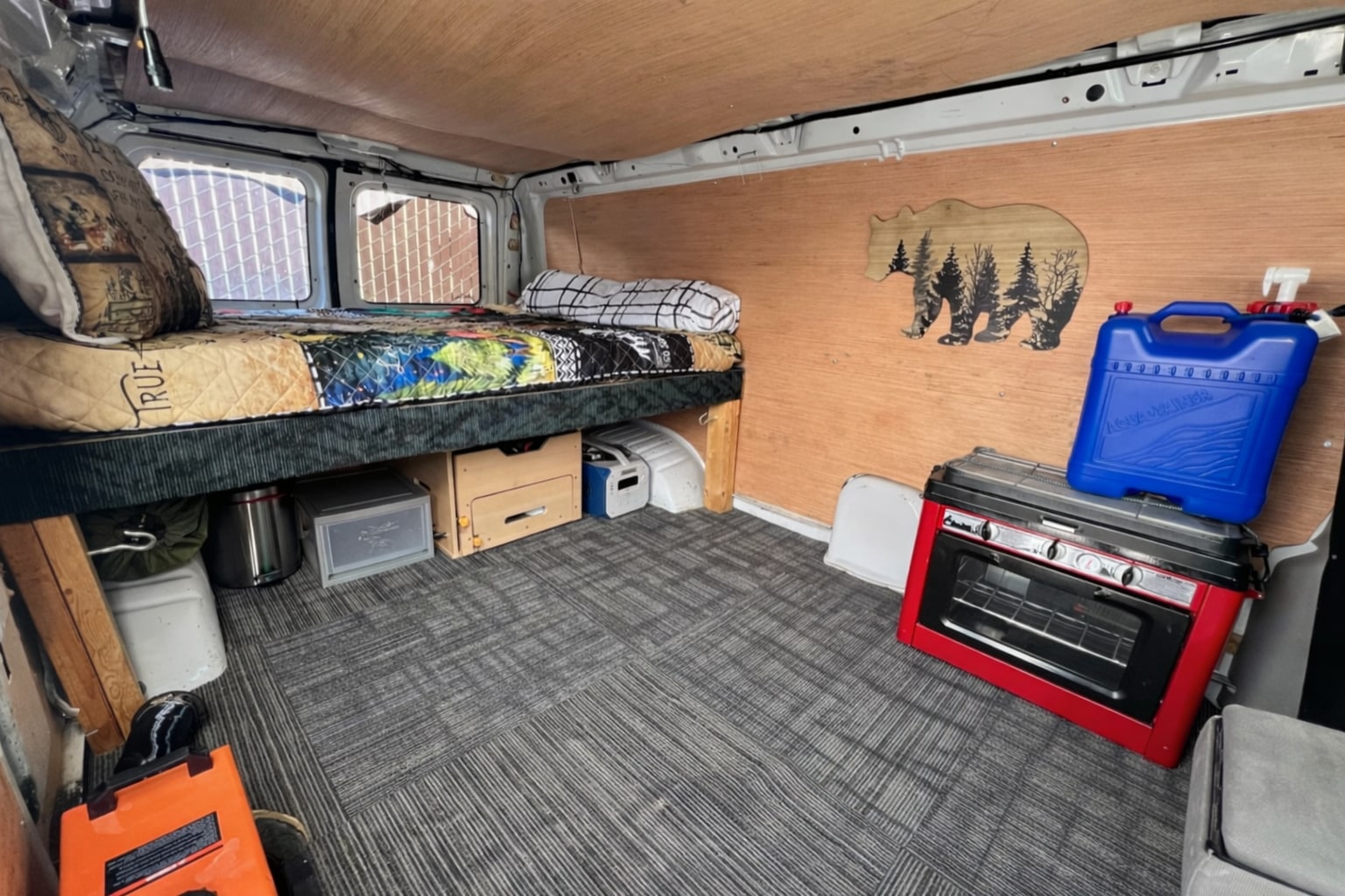 RV rental interior: cozy bed with under-bed storage, red portable stove, blue water jug, and a wooden bear decoration.. Ford Econoline 150 1997