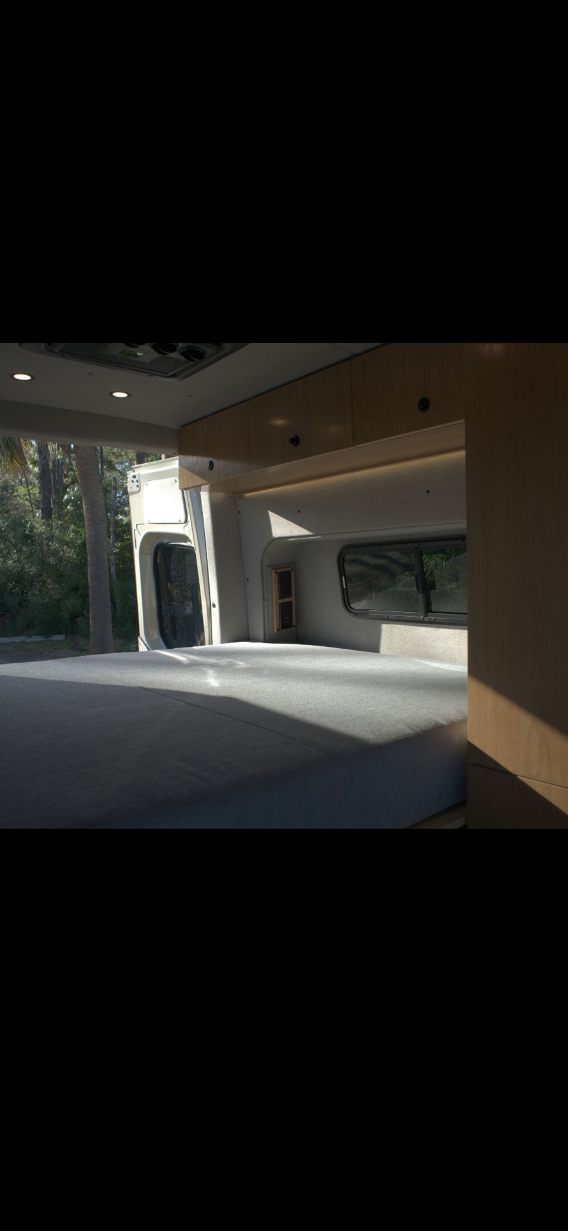 Cozy RV rental interior with a spacious bed, wooden cabinets, and a window revealing a serene outdoor nature scene.. Mercedes Motorhome 2025