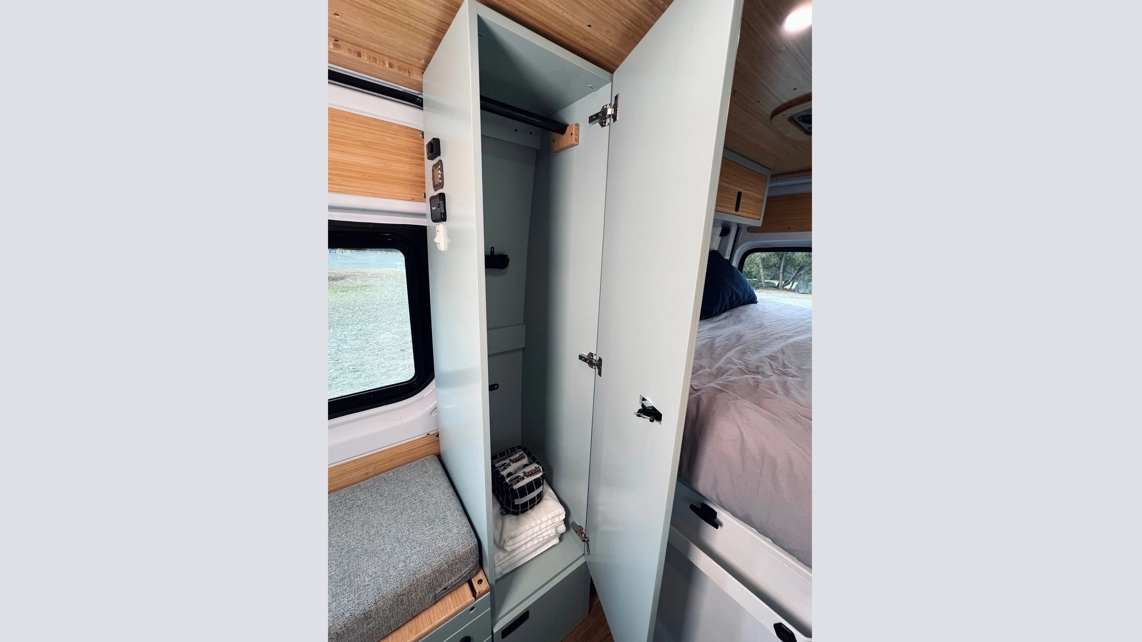 RV rental van interior features an open closet with towels, a bed, and a window view. Includes storage and seating.. Mercedes-Benz Sprinter 2023