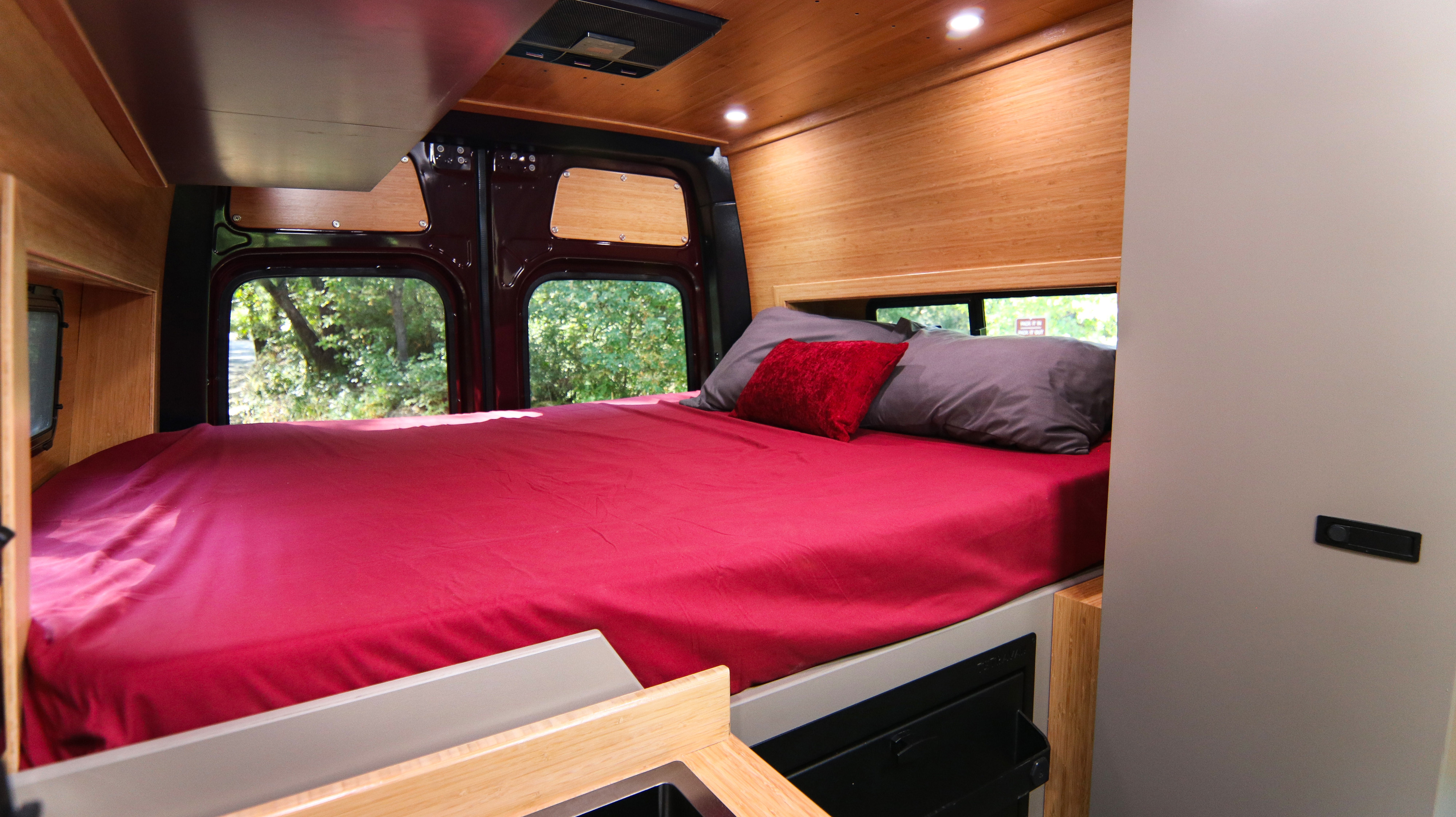 An inviting RV rental bed with red sheets & soft pillows, set in a wood-paneled camper van interior with natural views.. Mercedes-Benz Sprinter 2025