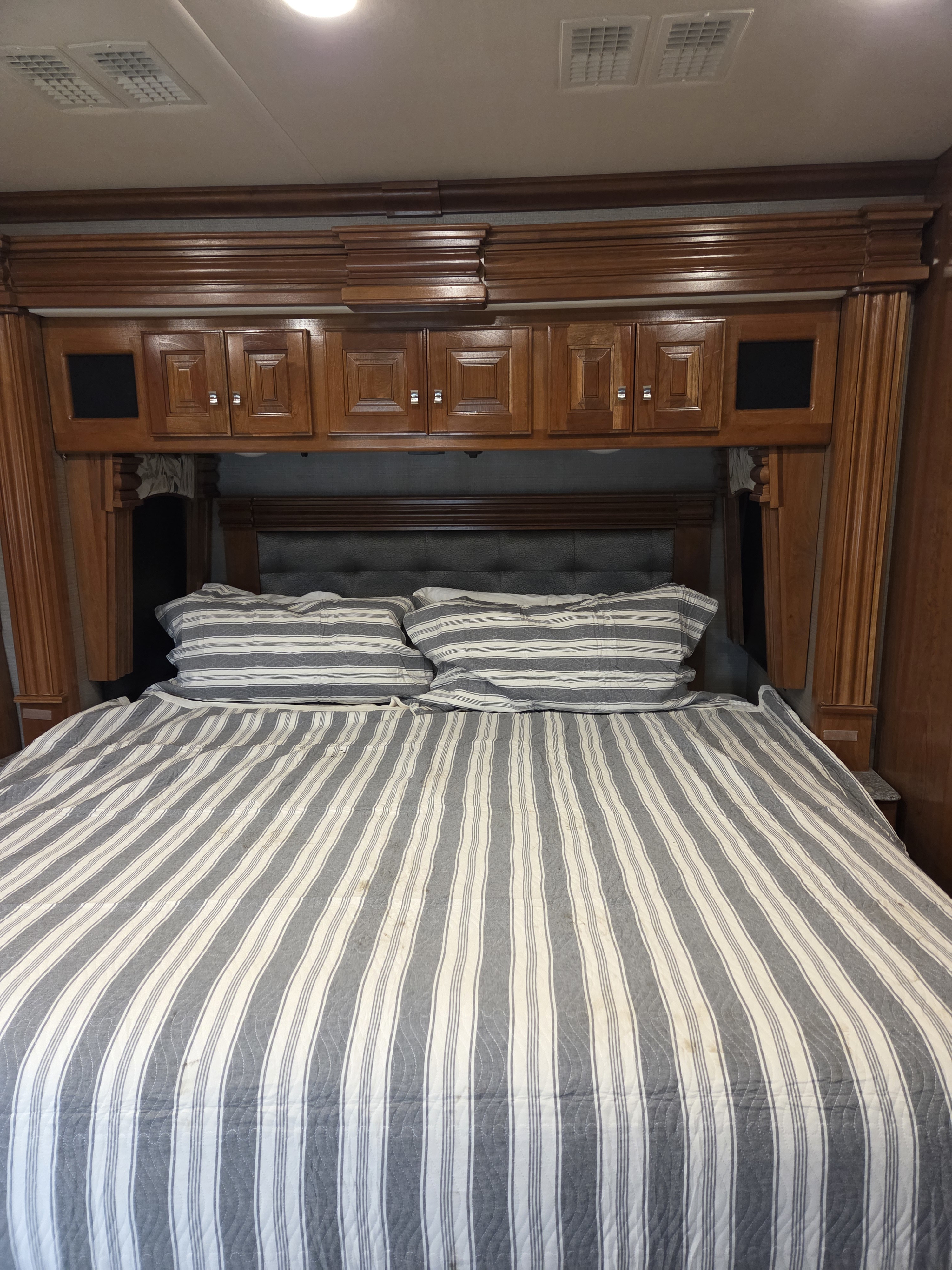 Spacious RV rental bedroom featuring a comfy bed with striped bedding, tufted headboard, wood cabinets & overhead AC vents.. Fleetwood Discovery LXE 2018